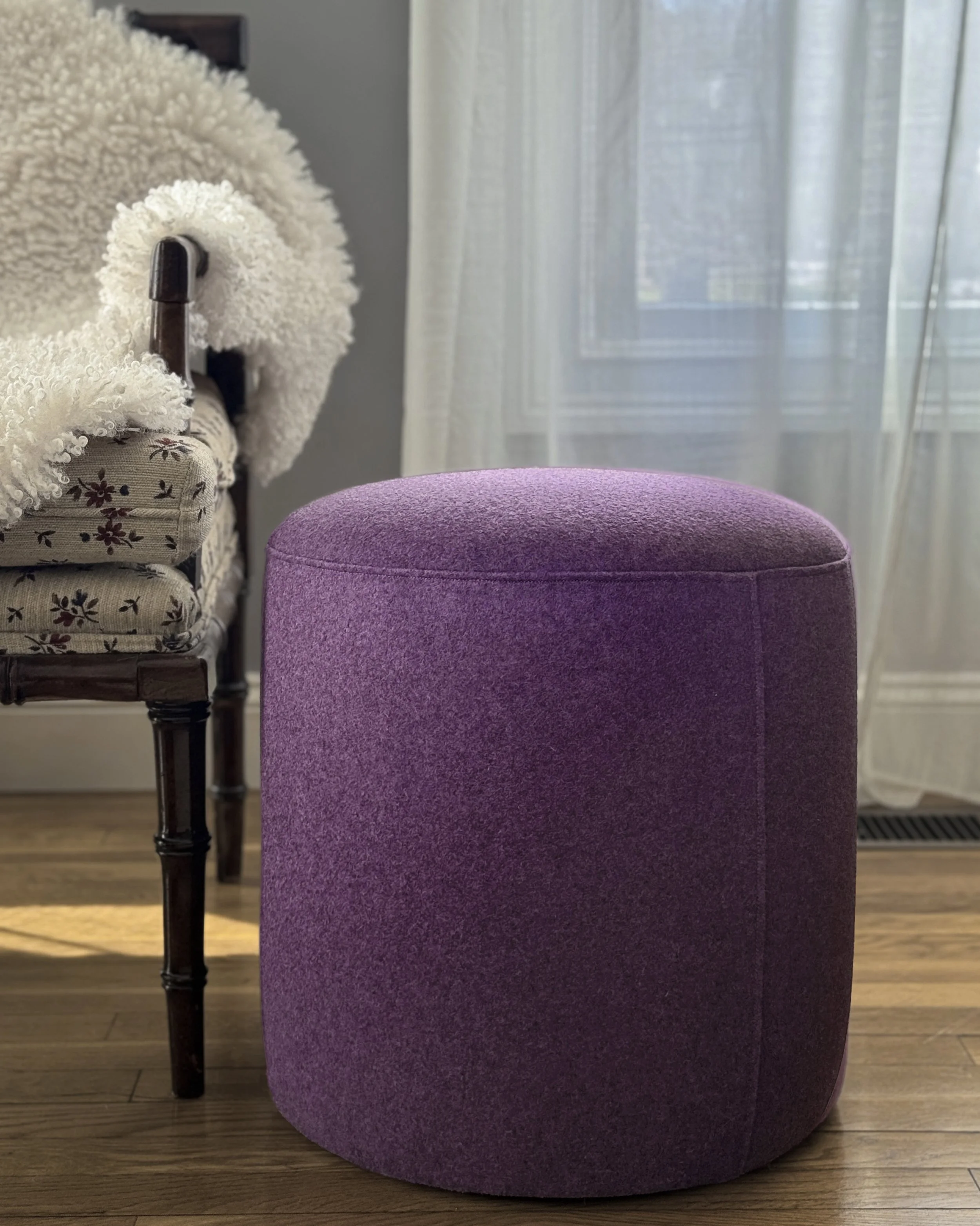 A custom, handcrafted purple upholstered ottoman in front of a beige floral patterned armchair with dark wood legs, situated on a wooden floor next to a window with sheer curtains.