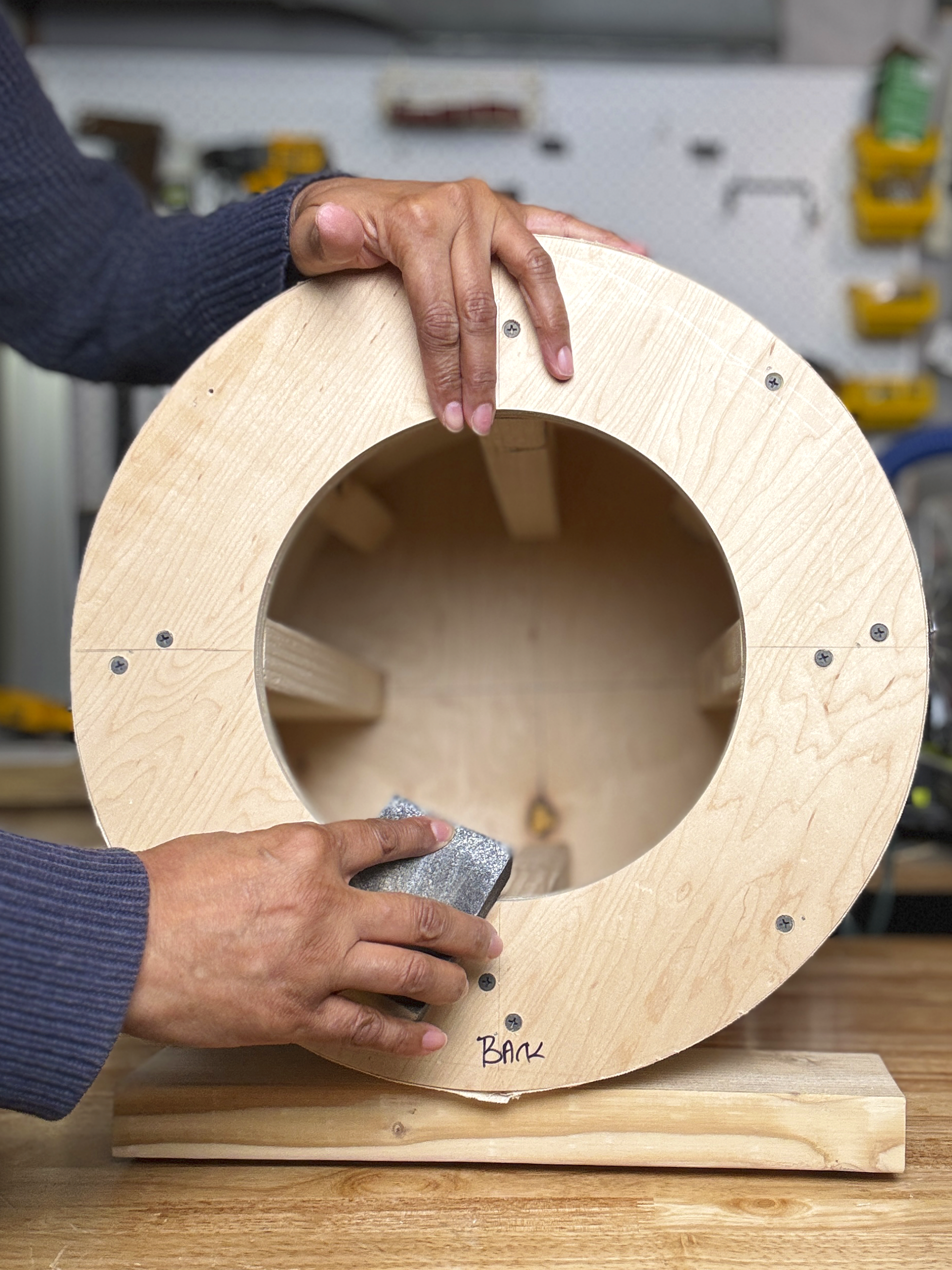An upholsterer building a custom ottoman out of wood. The upholsterer is using a sanding tool on the wood, and the background features a workshop with tools on a pegboard.