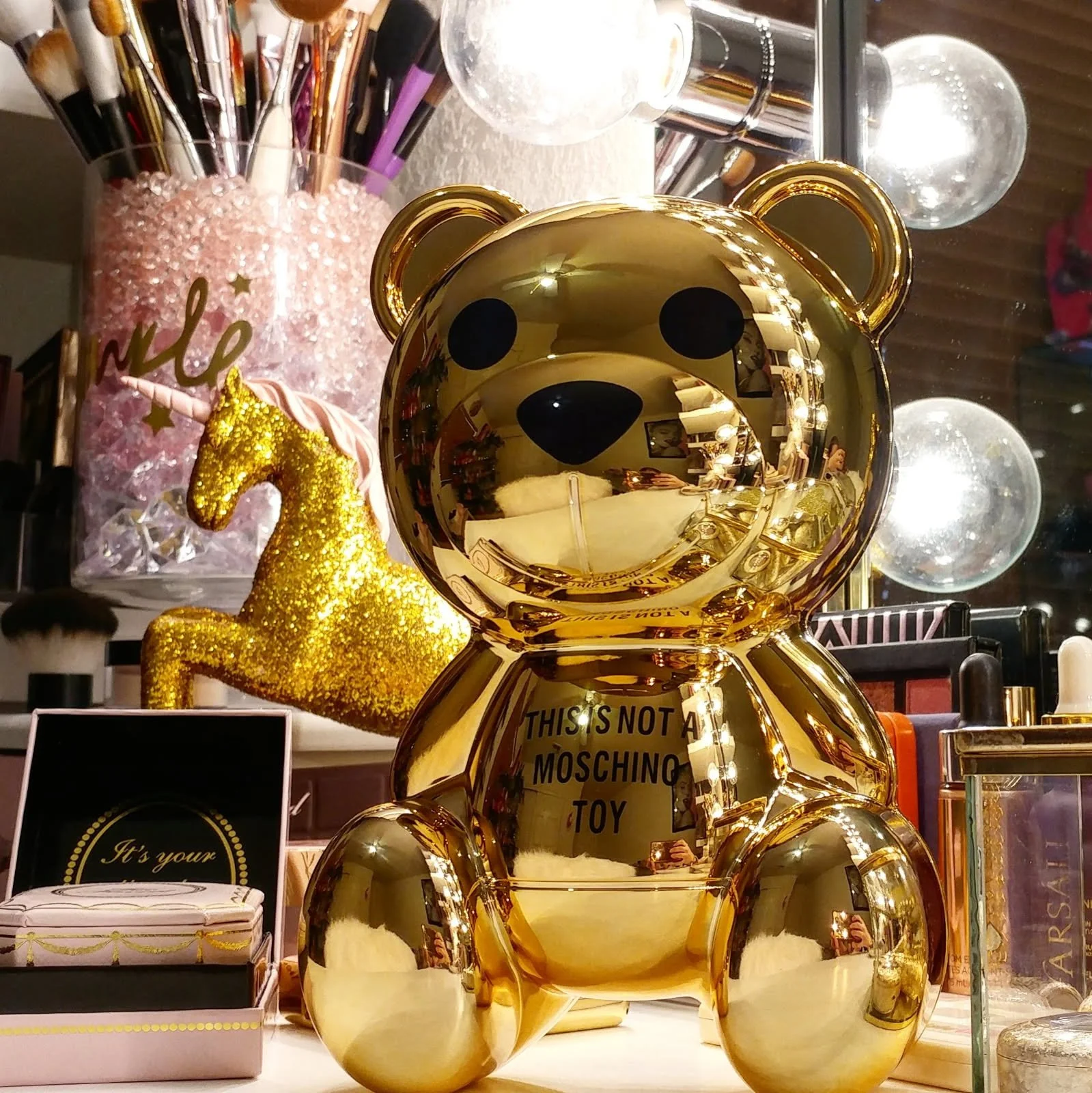 Golden bear-shaped decorative item with a reflective surface, placed on a vanity surrounded by makeup products, a glittery gold unicorn statue, and a pink container with makeup brushes.