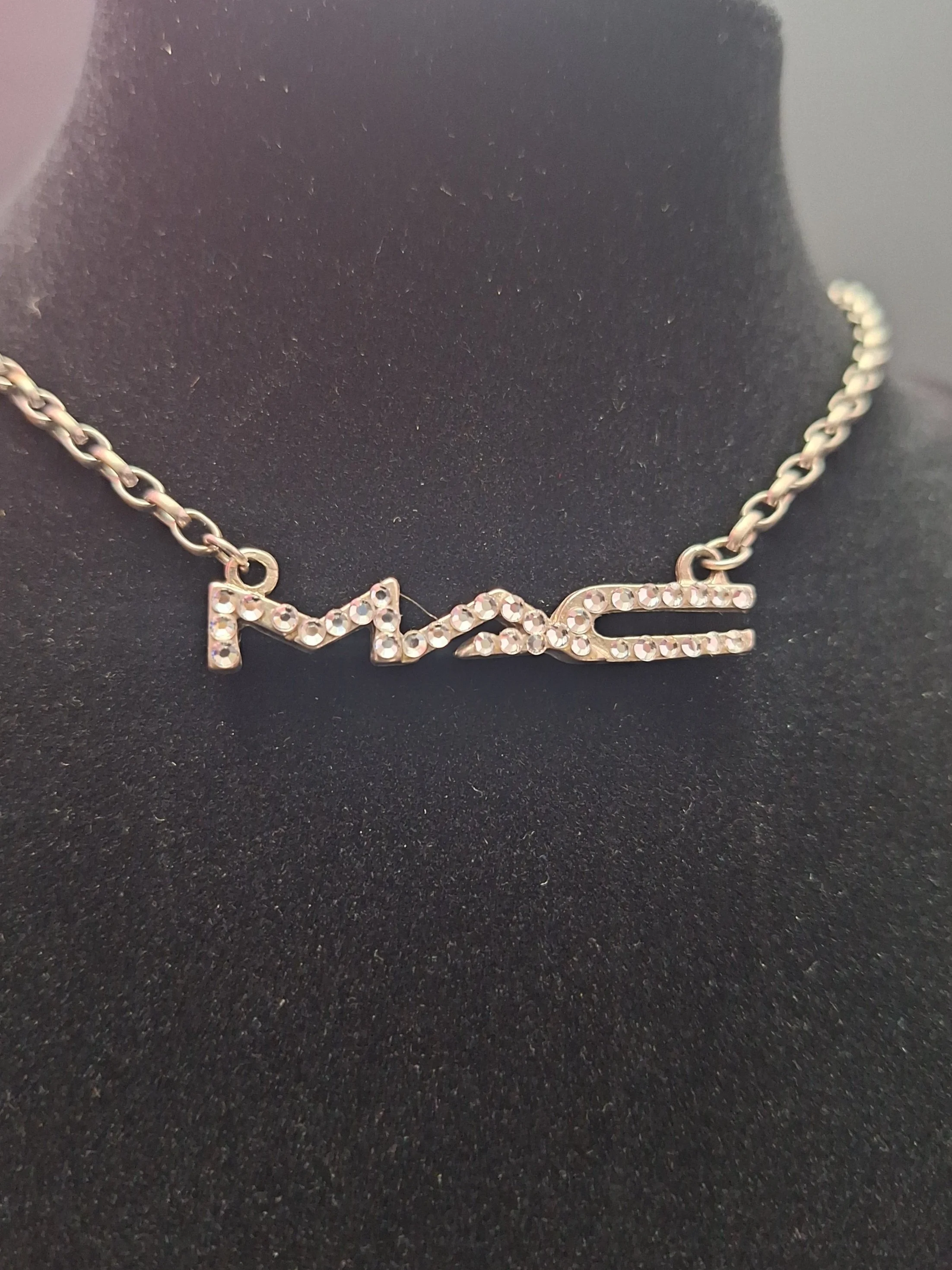 A silver necklace with the word 'MAC' spelled out in rhinestones, displayed on a black jewelry bust.
