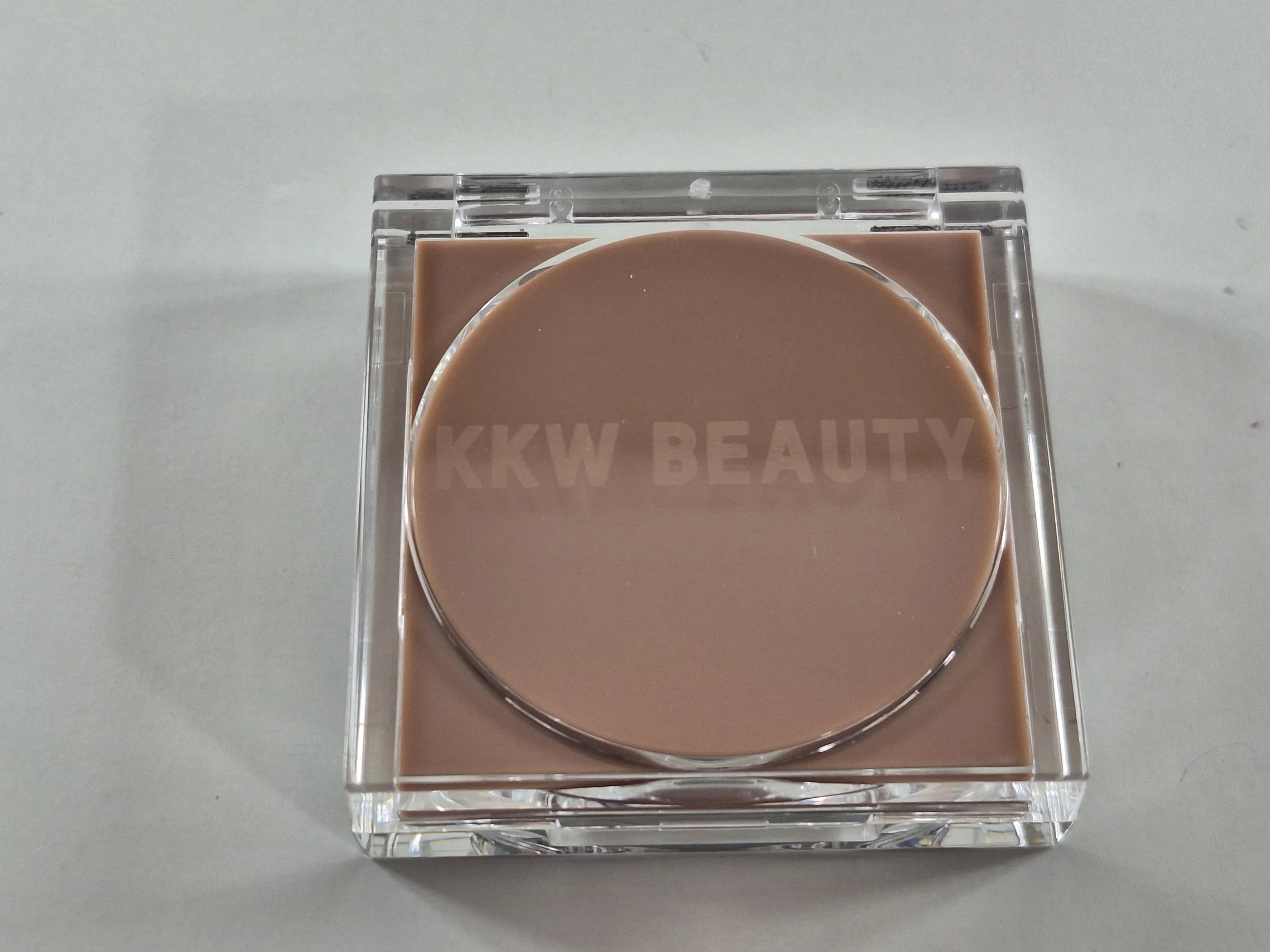 A square transparent plastic container with a round, brown cosmetic product and the text "KKW BEAUTY" embossed on the surface.