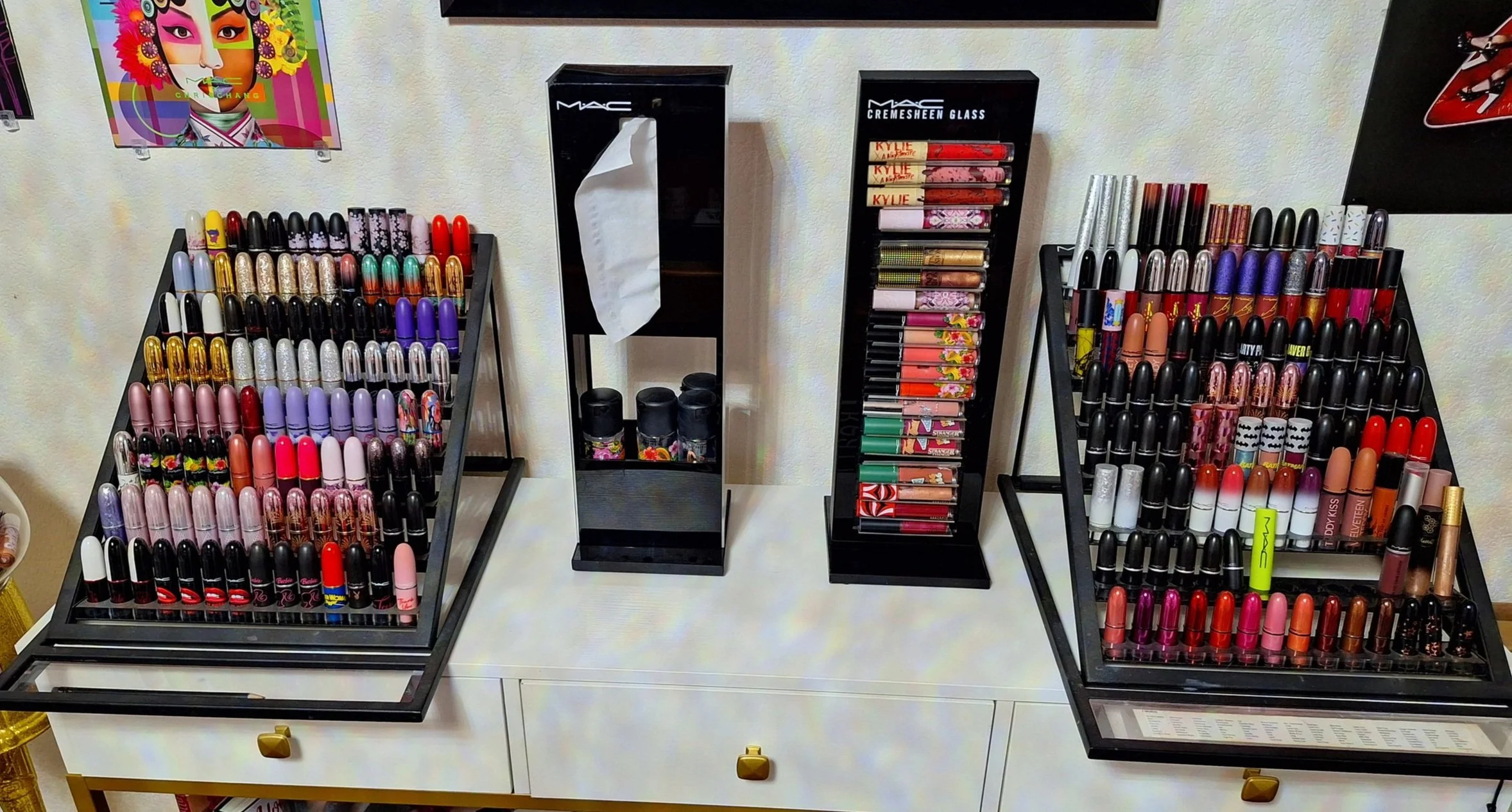 Display of various lipsticks and lip glosses arranged on black tiered stands on a white table, with colorful posters on the wall in the background.