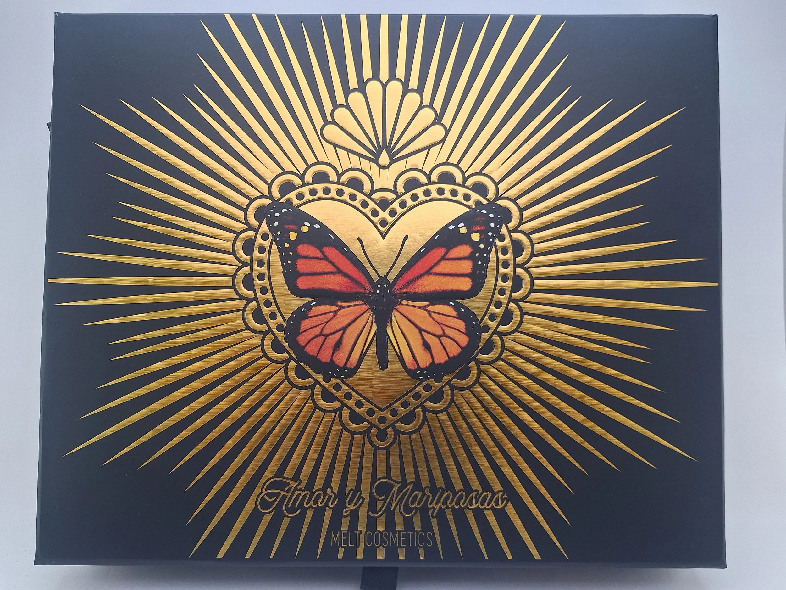 Black box with gold rays emanating from a central heart shape. Inside the heart, there's an orange and black butterfly. The text at the bottom reads 'Amor y Mariposas Melt Cosmetics'.