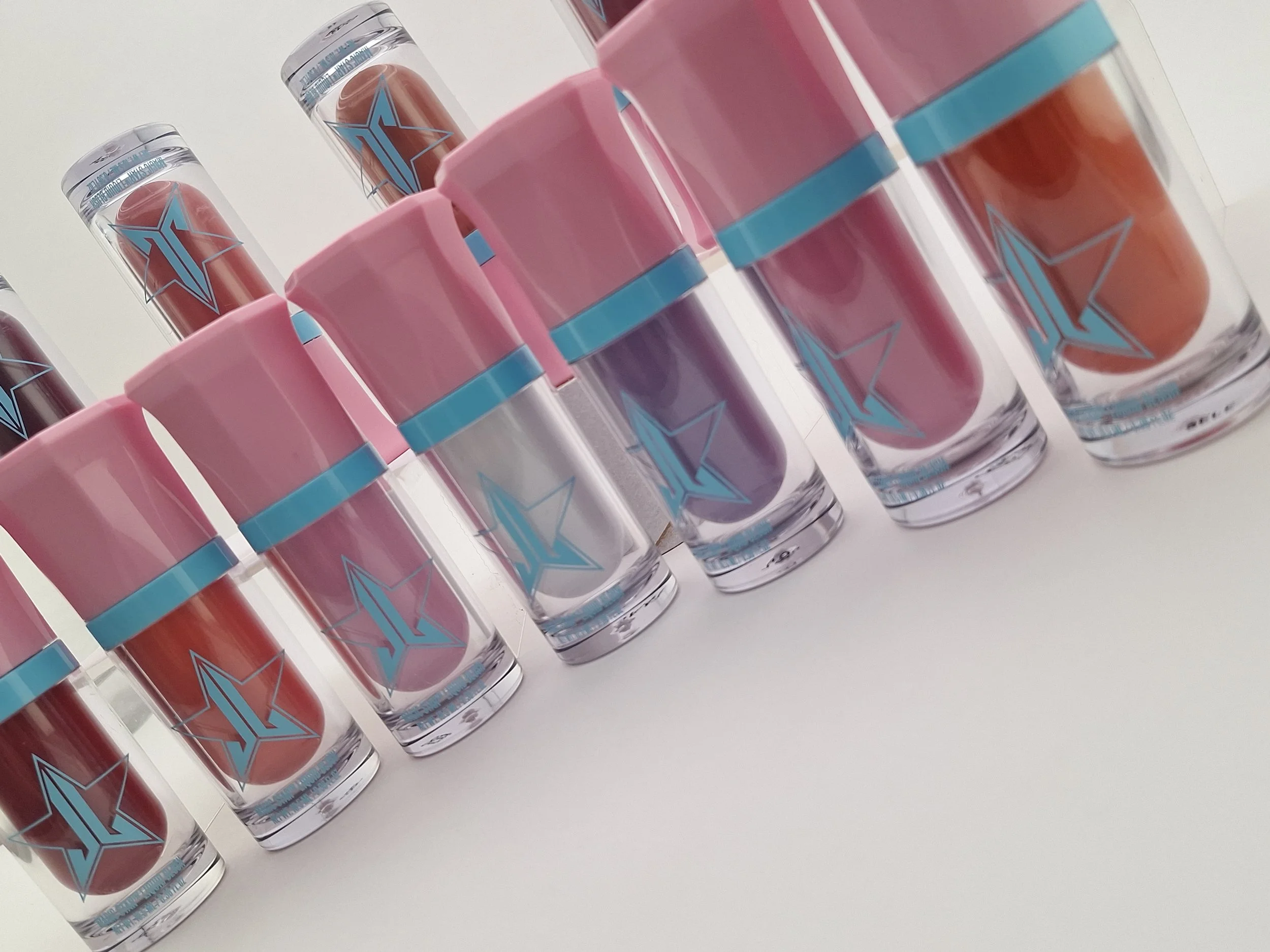 Multiple tubes of lipstick with pink and purple shades, each tube has a clear body with a pink cap and a blue star logo on the front, arranged in a row on a white surface.