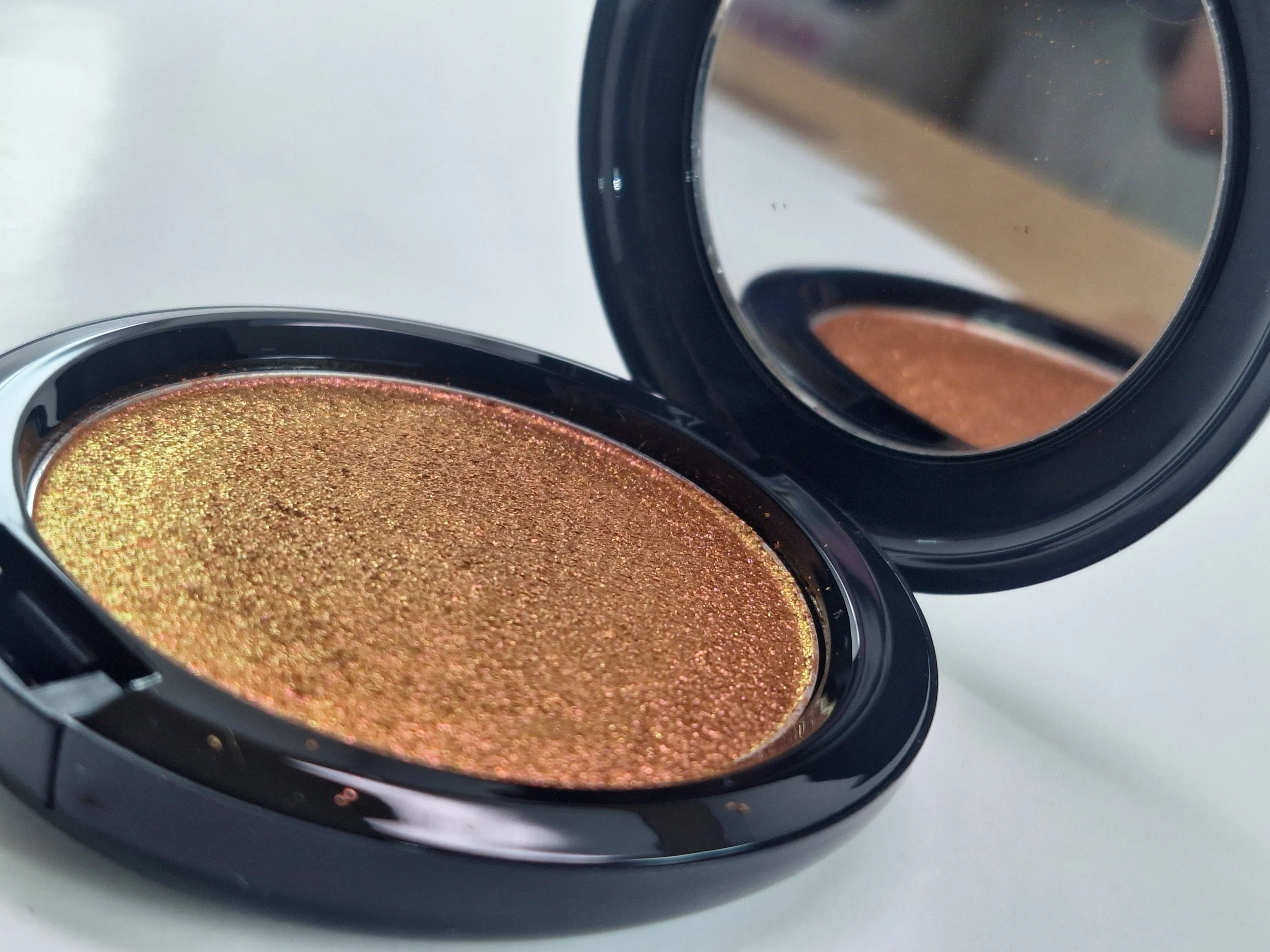 Close-up of a shimmering gold makeup eyeshadow in a black compact case with an attached mirror.