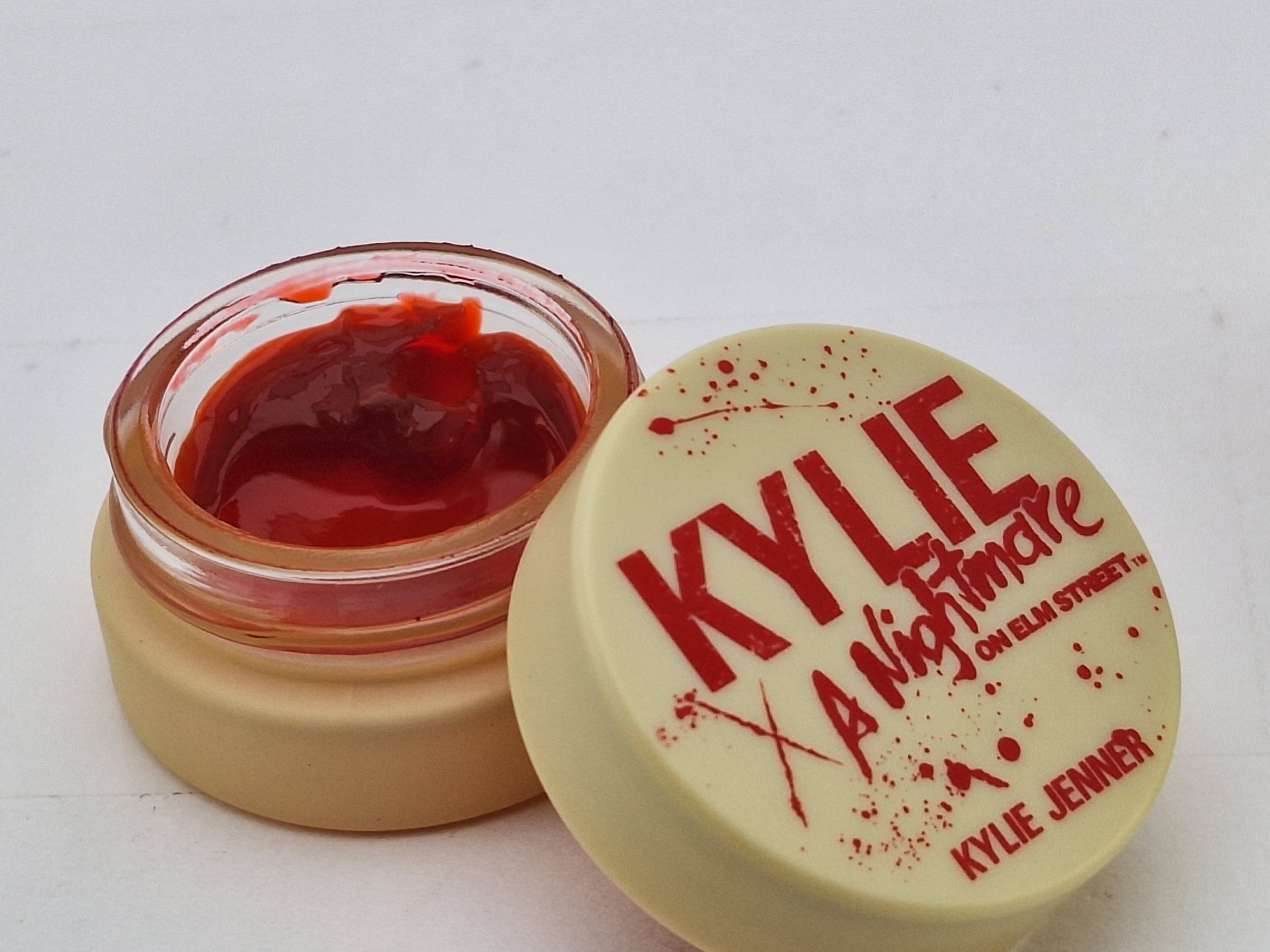 Open jar of red lip gloss with the lid beside it, featuring the words 'KYLIE' and 'A NIGHTMARE' in red, with splatter design and 'KYLIE JENNER' written at the bottom.