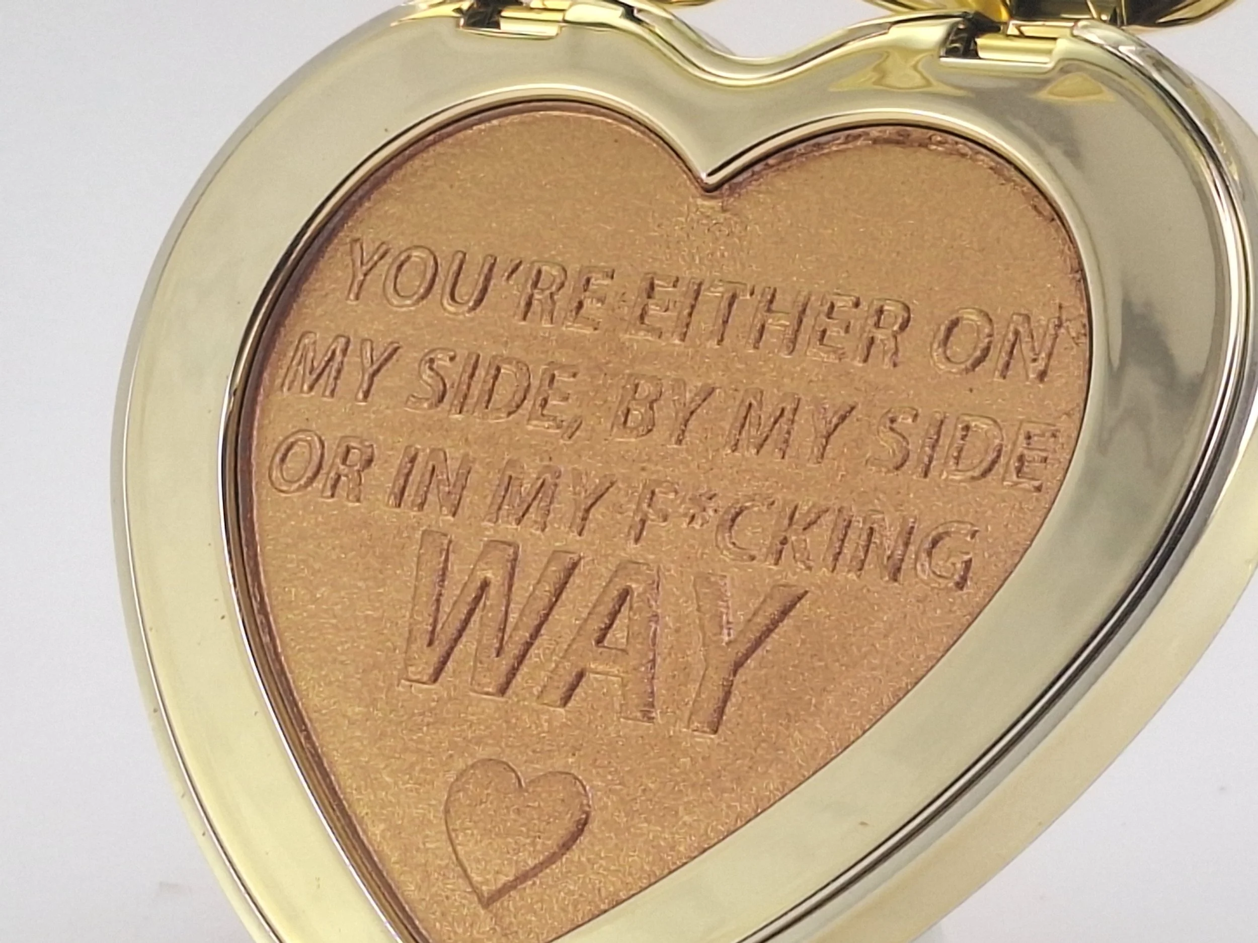 A heart-shaped compact makeup with a gold rim, containing bronzer with an engraved message: "You're either on my side, by my side or in my f*cking way" and a small heart illustration.