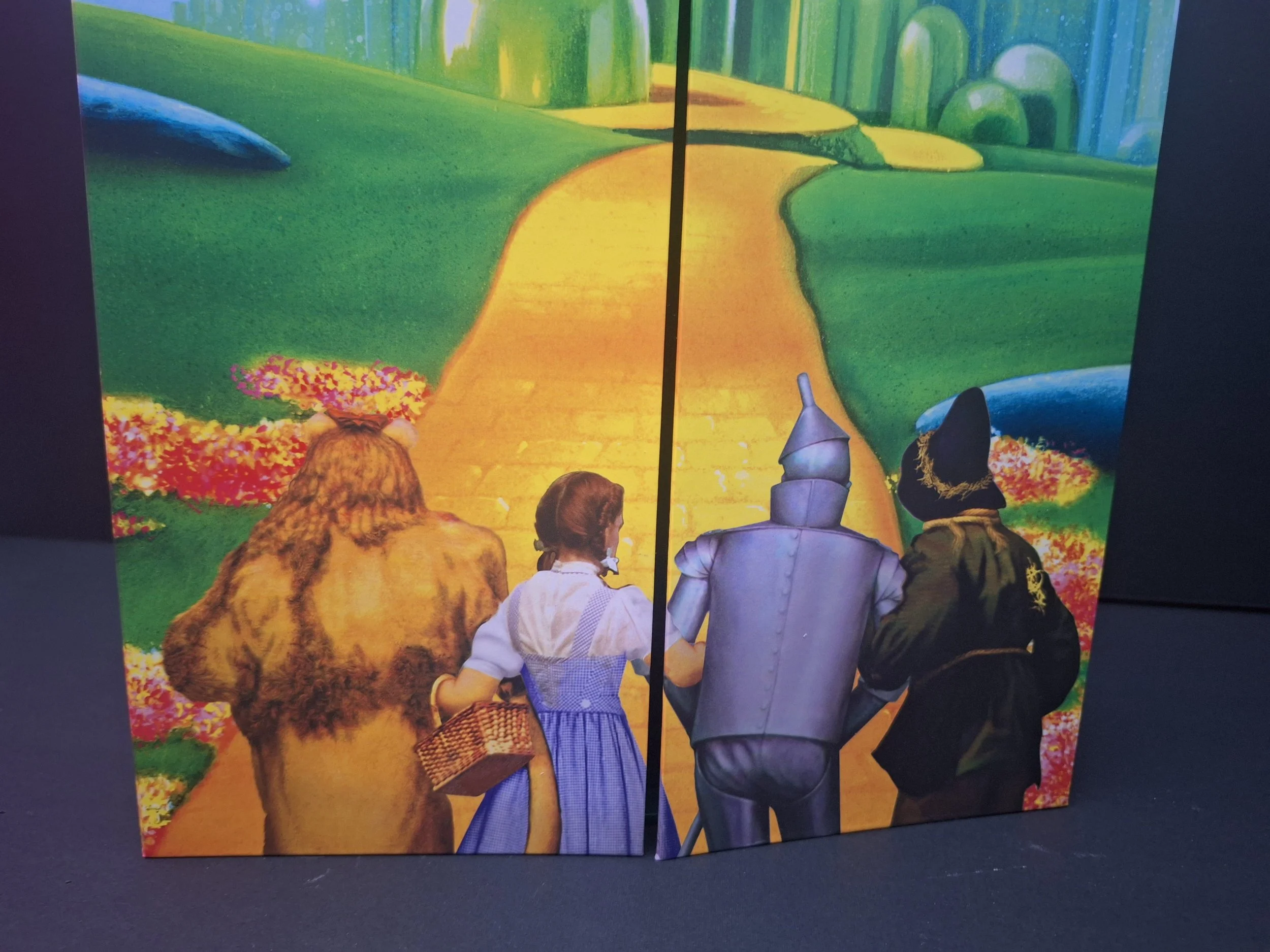 Colorful illustration of four characters—lion, girl in dress, tin man, and witch—walking down a yellow brick road through a vibrant green landscape with flowers and trees.
