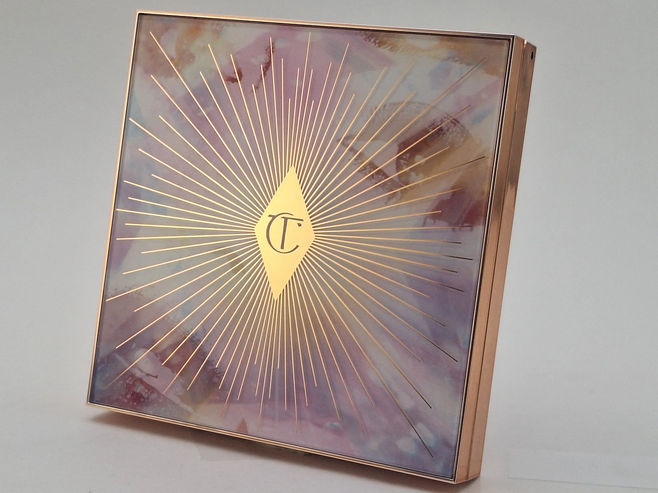 A square makeup palette with a shiny rose gold edge, featuring a decorative starburst design with fine lines radiating from a central yellow diamond shape that contains the letter 'T' enclosed in an oval.
