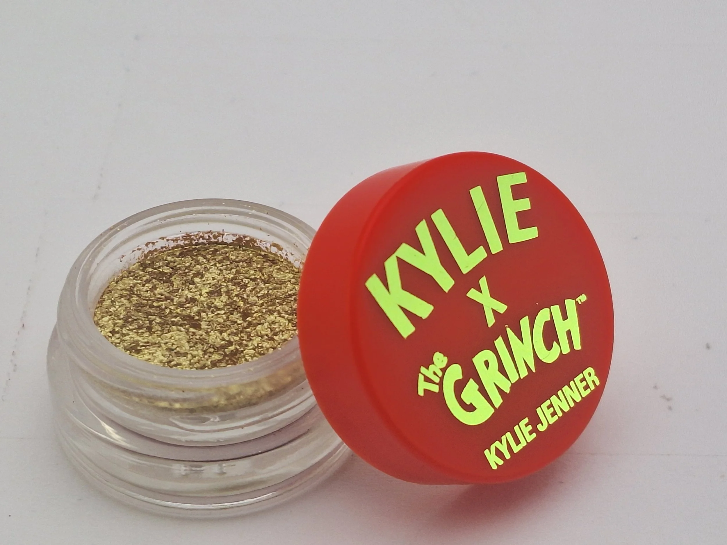 A small clear container filled with gold-colored glitter, placed next to a red round container lid with bright yellow text reading 'KYLIE x THE GRINCH KYLIE JENNER'.