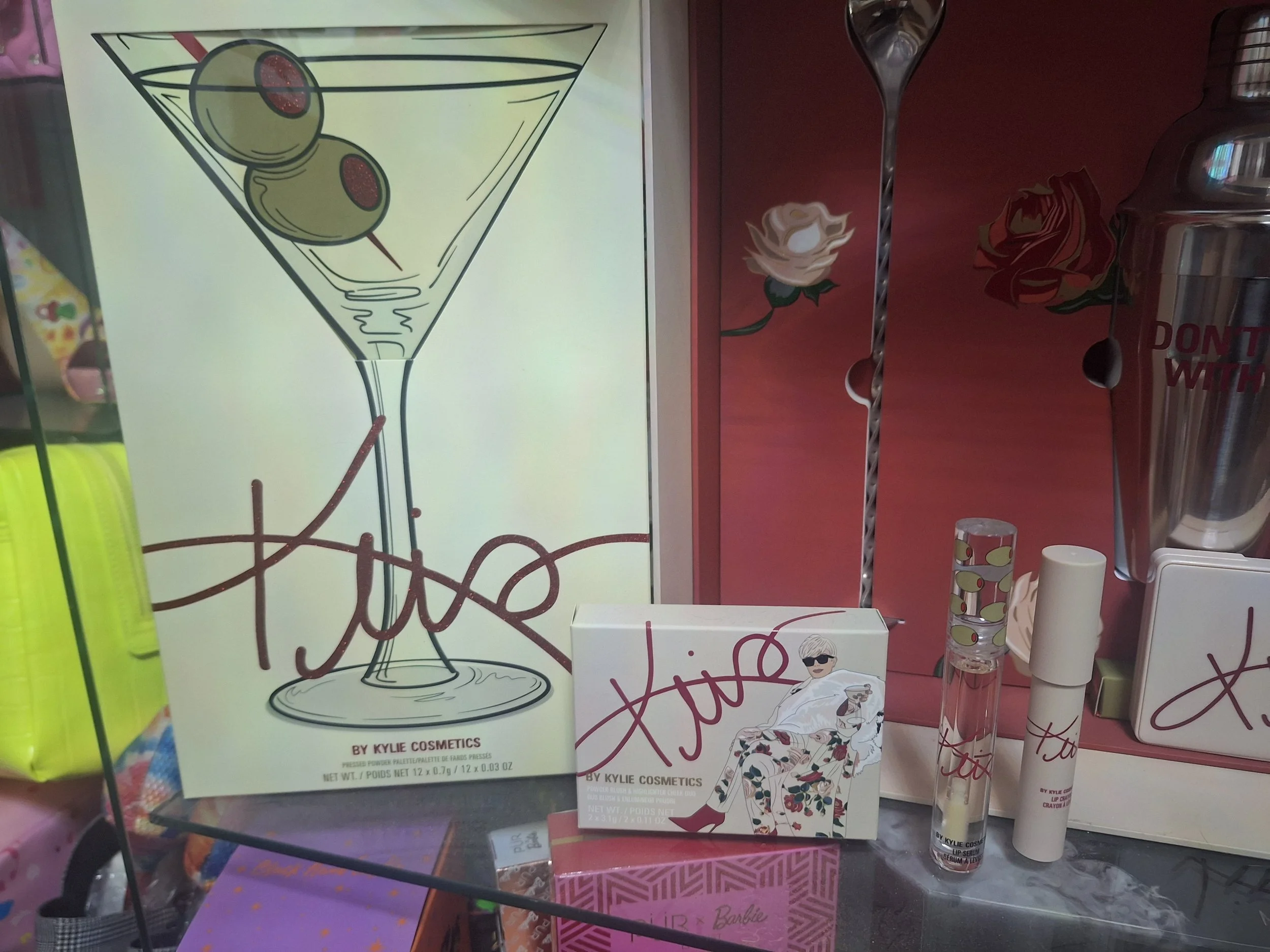 Kylie Cosmetics makeup display with poster, lip gloss, lipstick, and blush products. The poster shows a martini glass with olives and the signature Kylie logo.