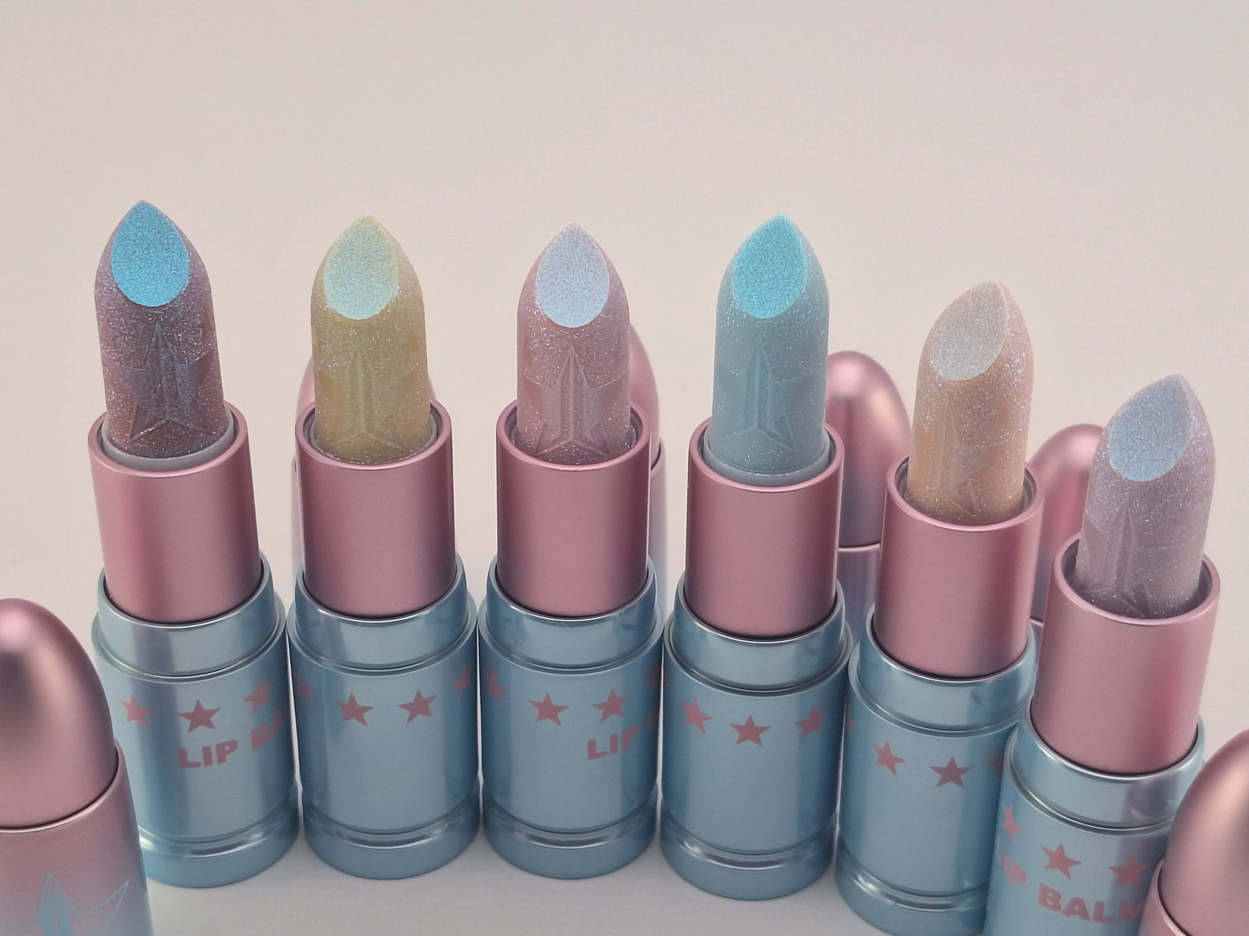 A collection of six pastel-colored lipstick tubes with glittery tips, arranged in a row on a white surface, with pink and blue packaging with star decorations.