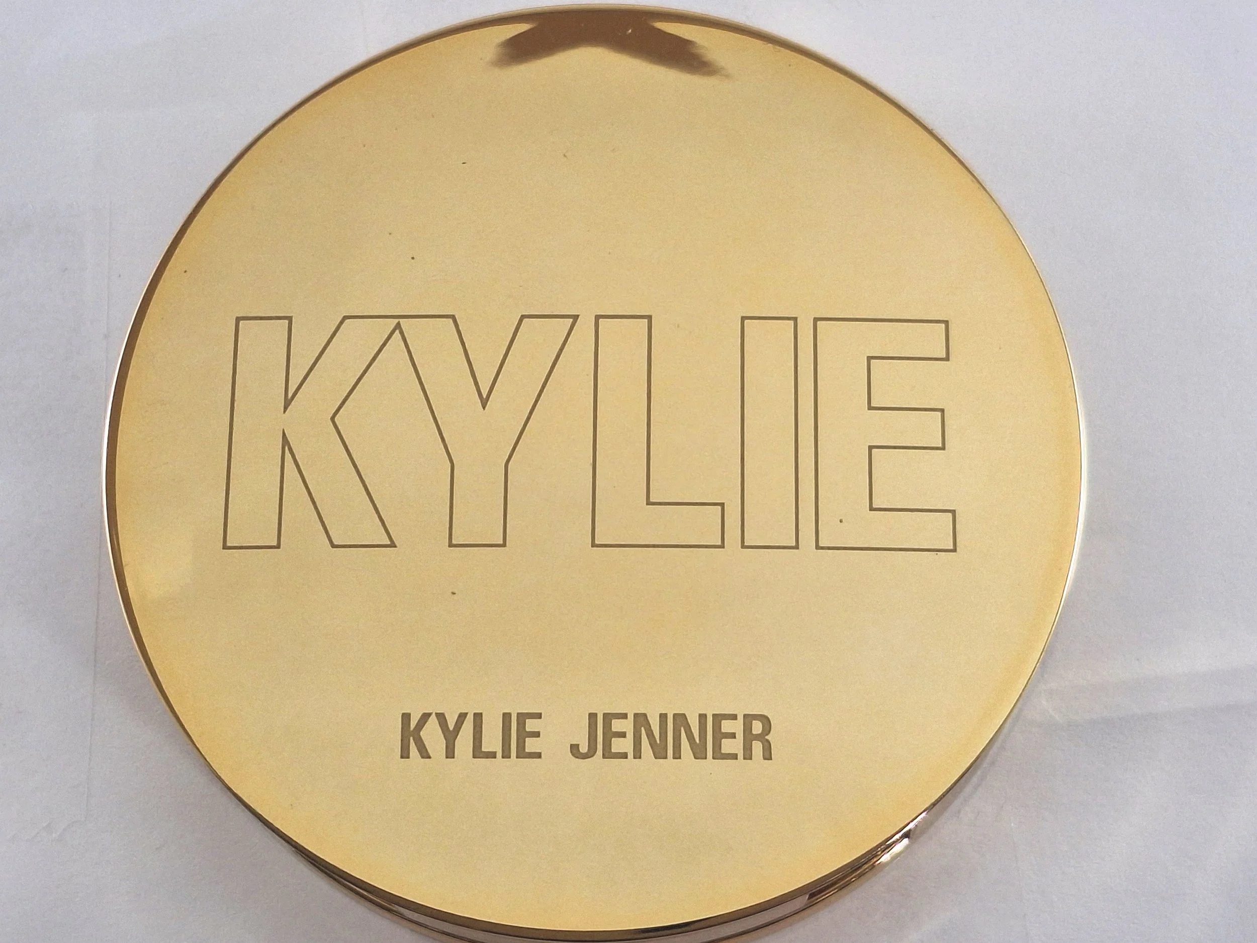 Golden compact mirror with the name 'KYLIE' outlined in large letters on the lid, and 'KYLIE JENNER' written below in smaller bold letters.
