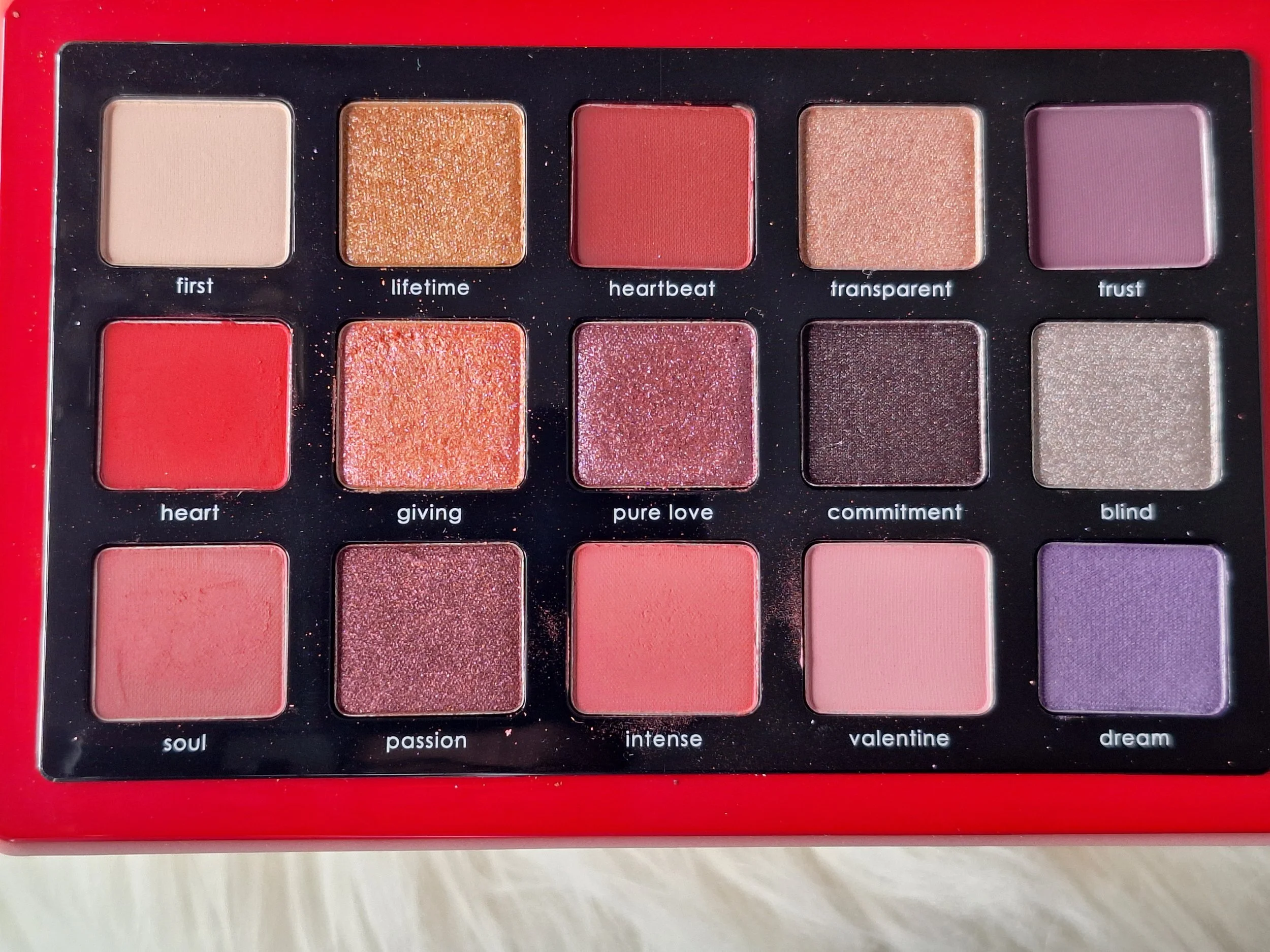 An eyeshadow palette with 15 shades arranged in a 3x5 grid, each labeled with a name like first, lifetime, heartbeat, transparent, trust, heart, giving, pure love, commitment, blind, soul, passion, intense, valentine, dream.