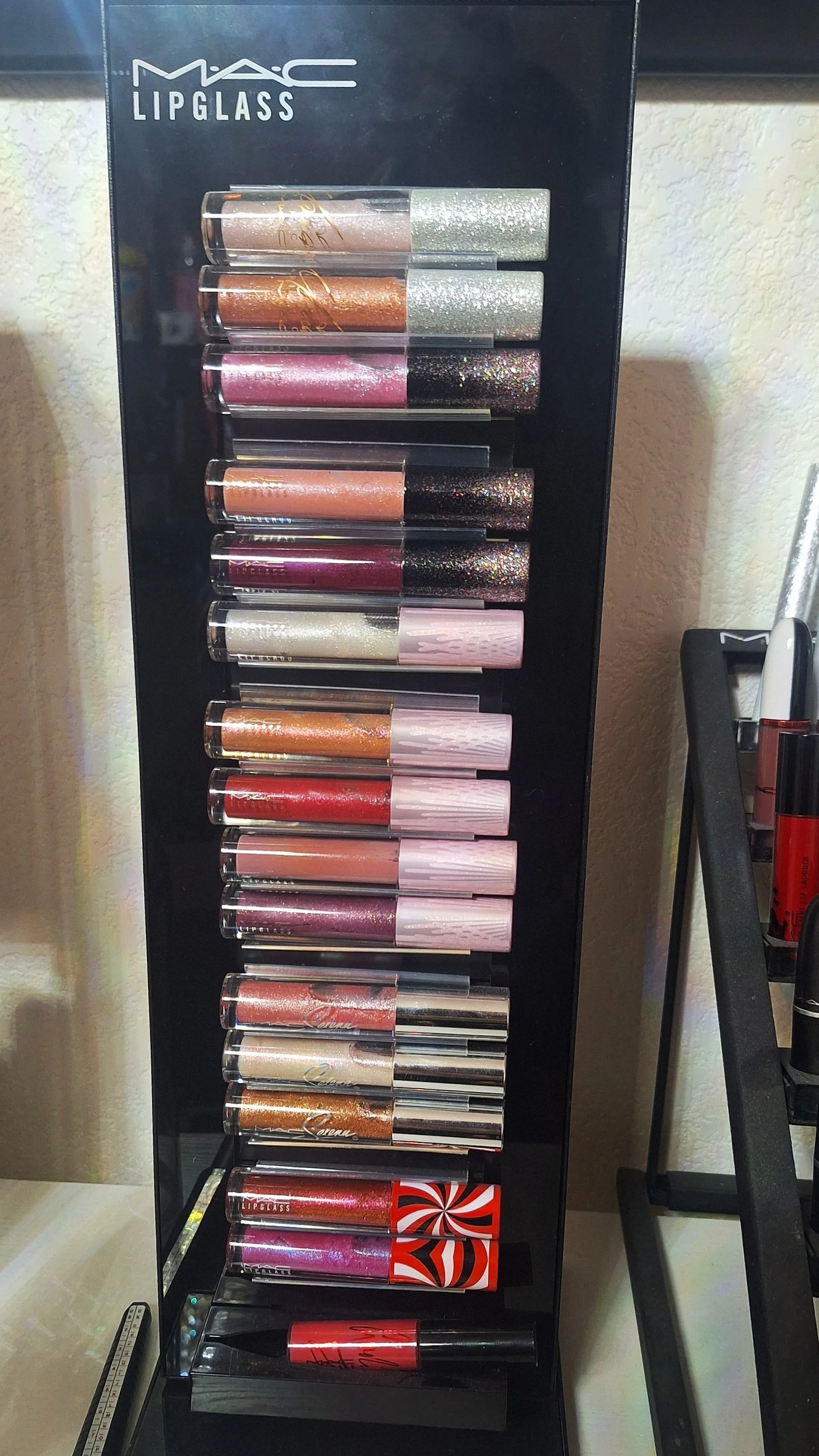 Display stand of MAC Lipglass with various lip glosses in glitter and metallic shades in multiple rows.