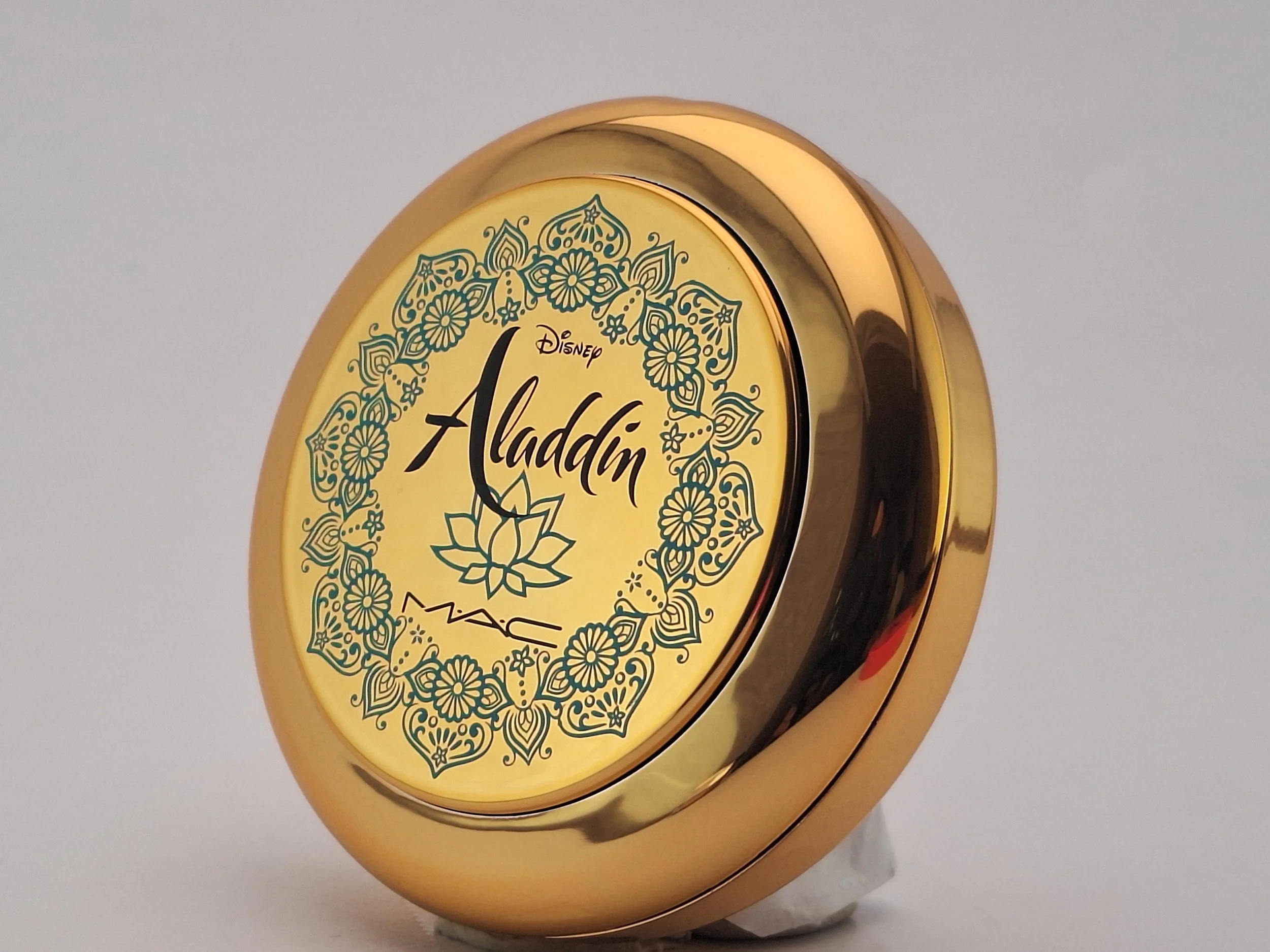 A round, gold-colored container with Disney's Aladdin logo and ornate floral design on the lid.
