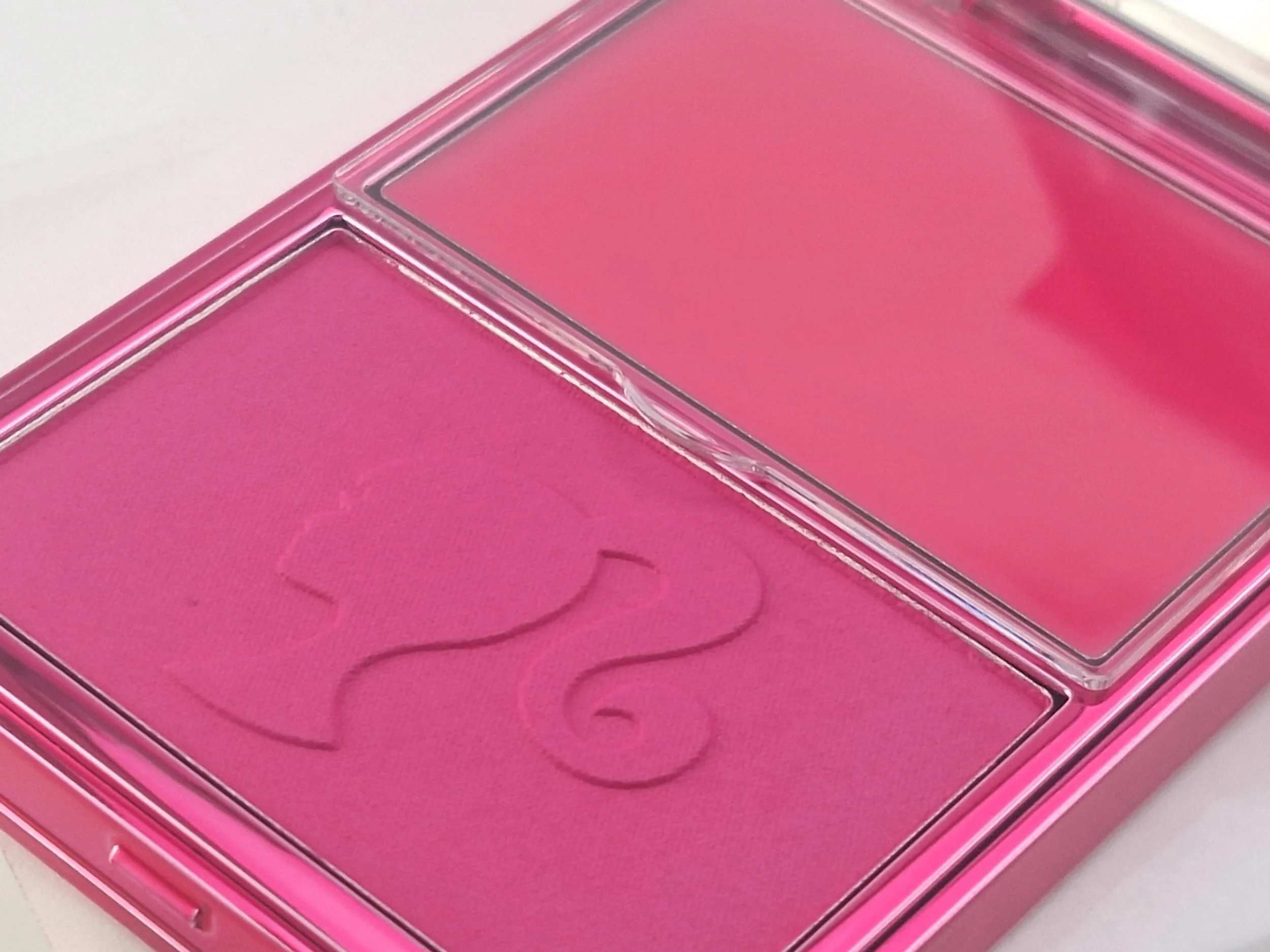 A pink makeup palette with two shades of pink blush, one with a floral imprint and one plain, in a transparent rectangle container.