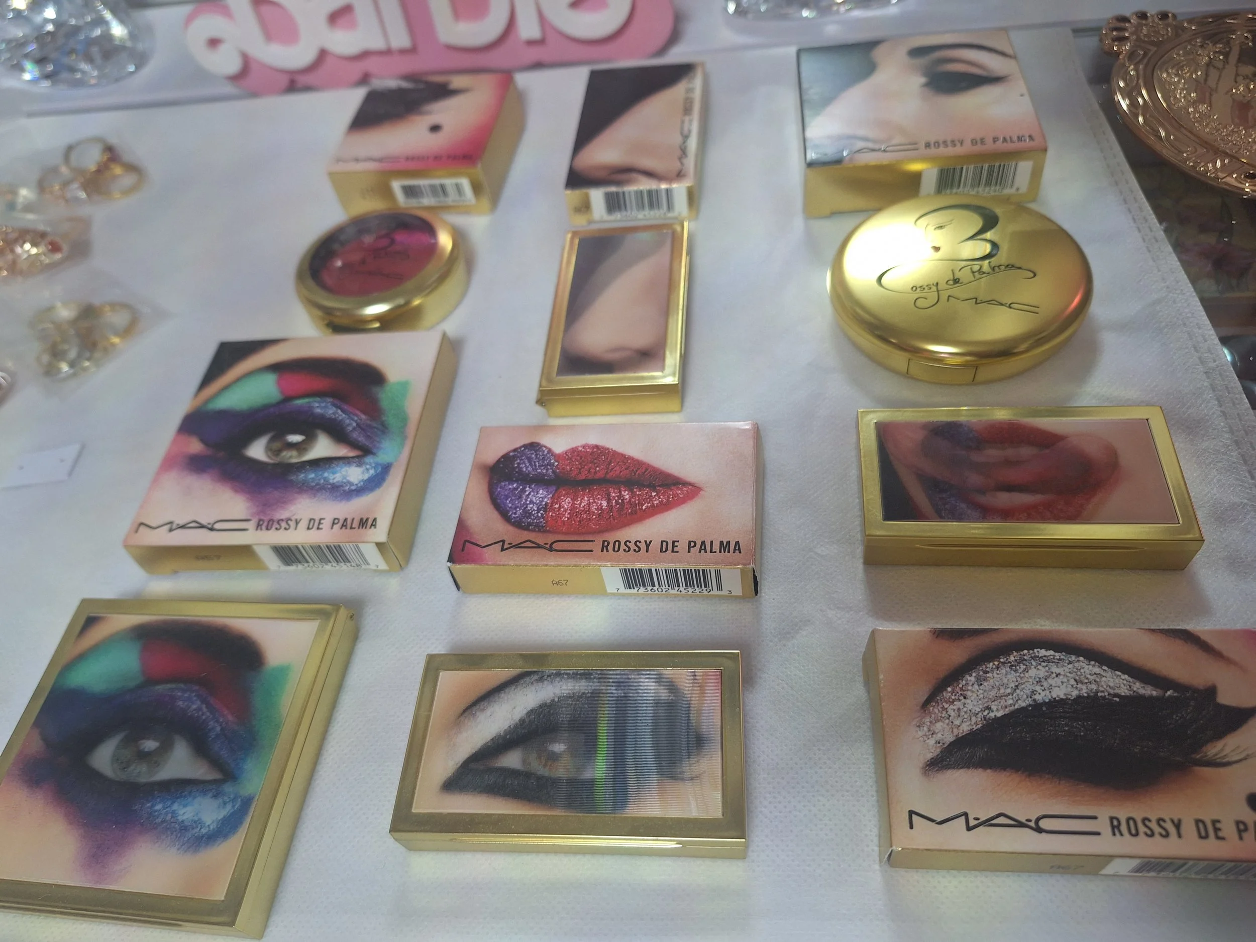 Makeup products displayed on a table, including eyeshadow palettes with colorful eye designs and lipstick boxes, surrounded by jewelry.