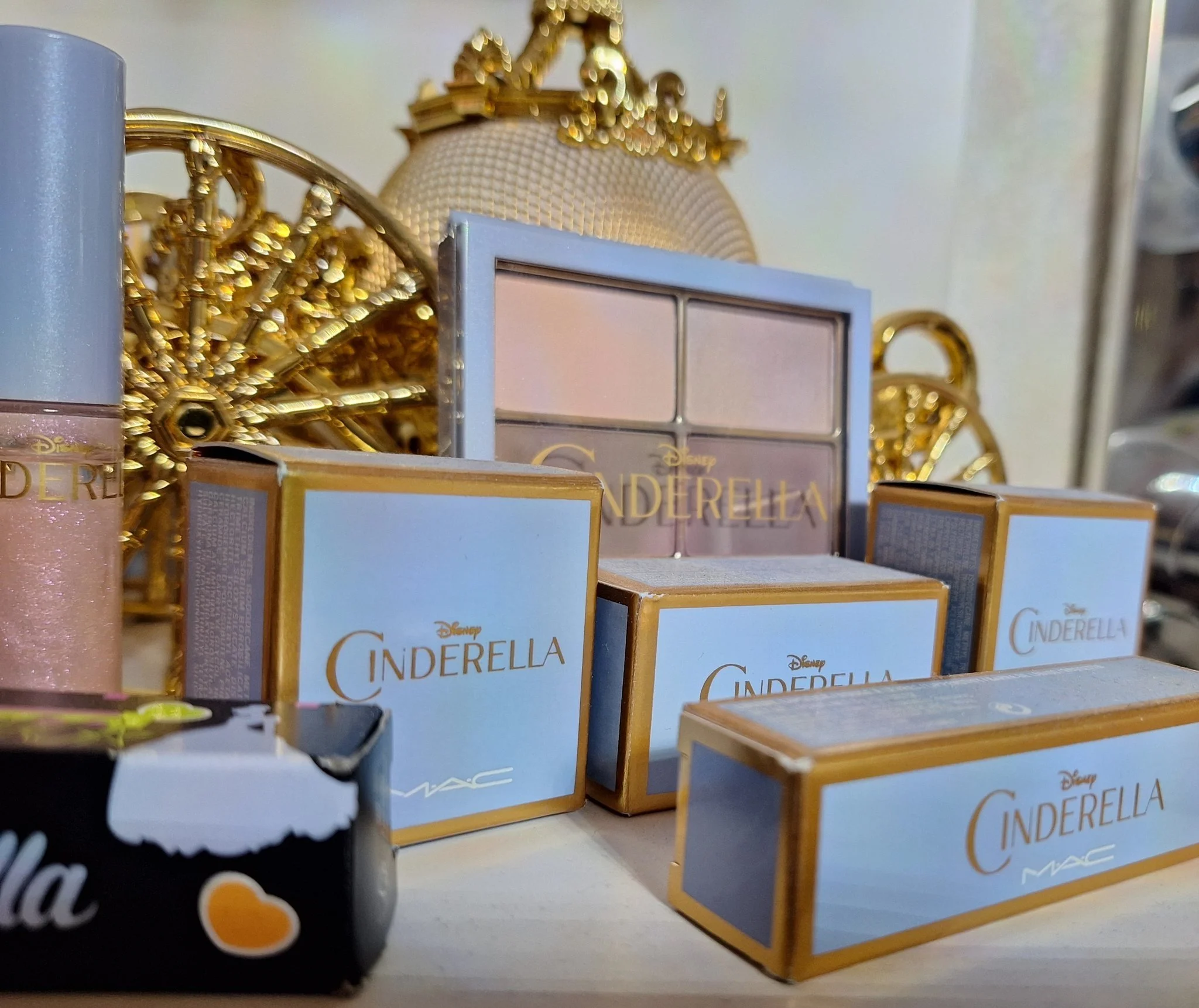 Various Cinderella-themed makeup products, including a palette and boxed items, displayed on a surface with decorative golden Cinderella figurines in the background.