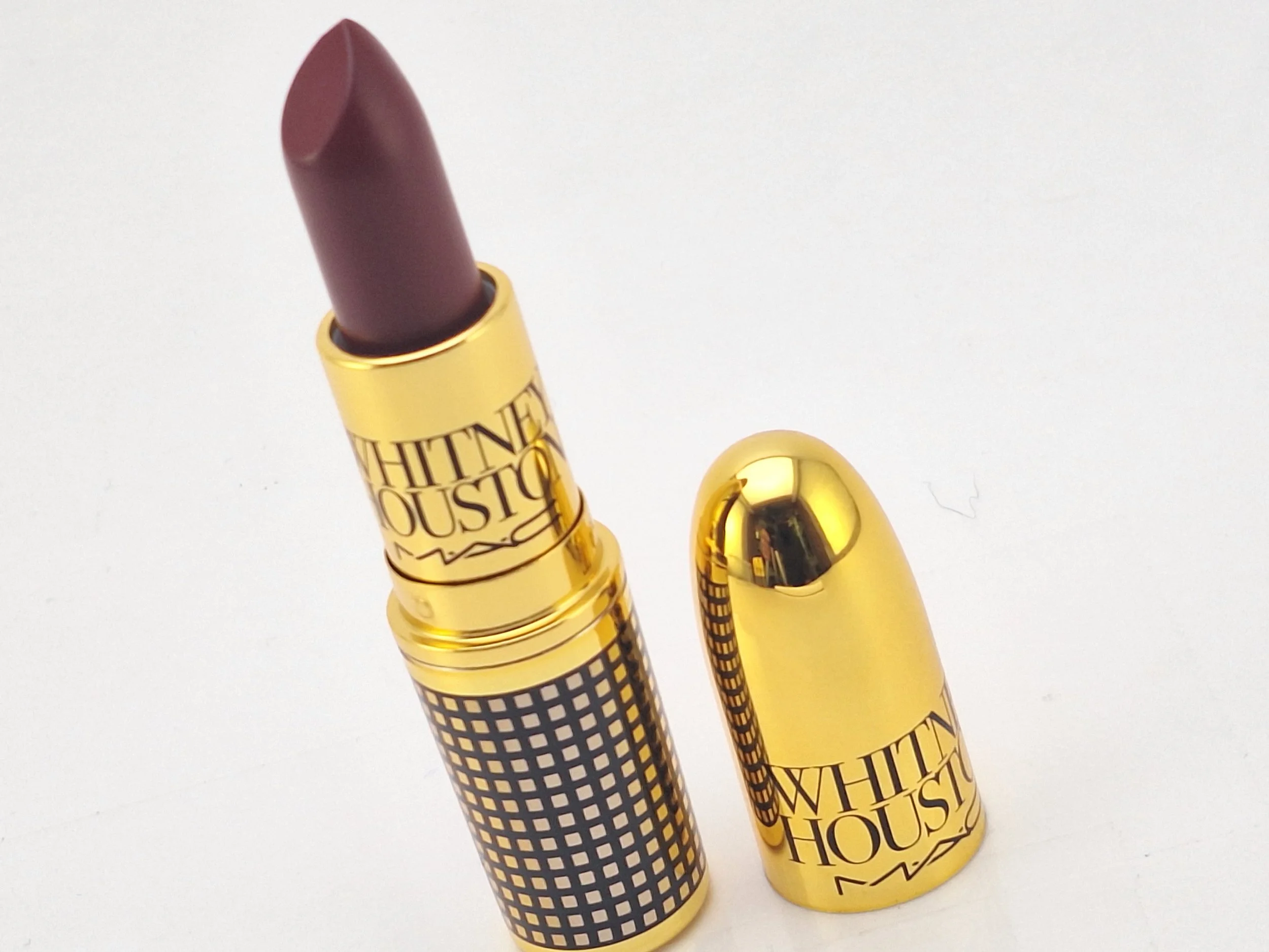 A MAC lipstick with a purple shade in a gold and checkered packaging, with the cap removed and placed beside it.