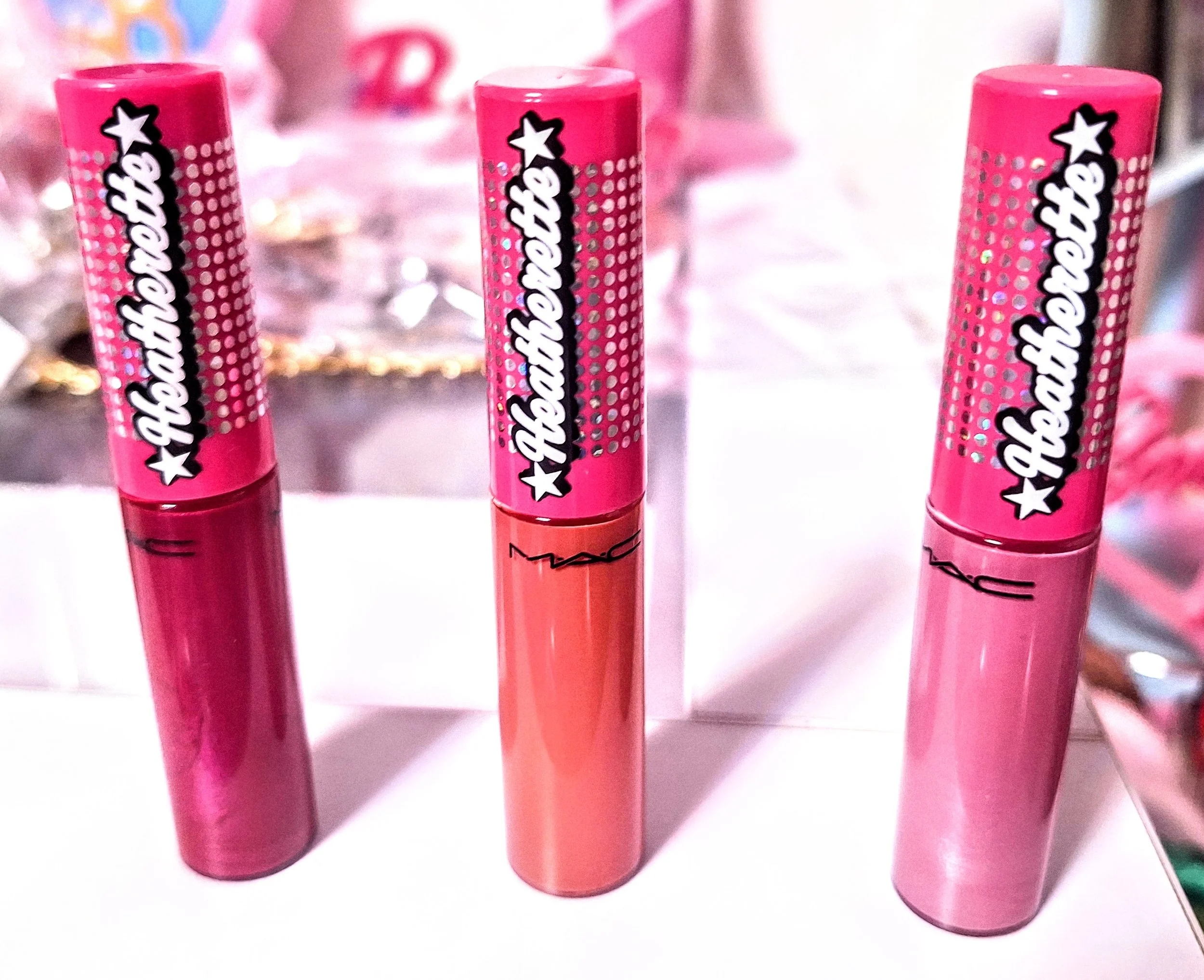Three MAC Heatherette lip glosses in bold pink, coral, and fuchsia with signature holographic packaging.
