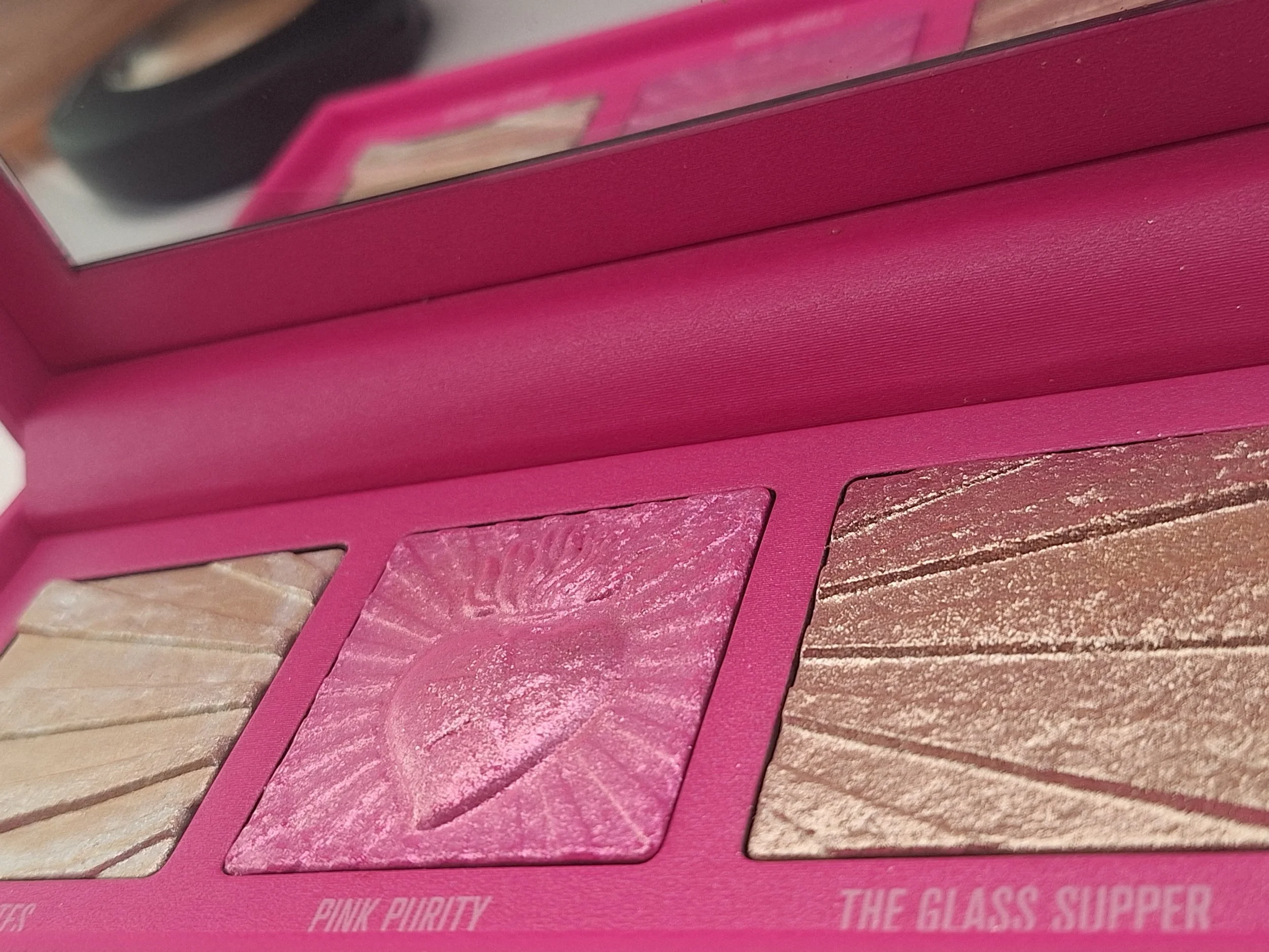 Close-up of an eyeshadow palette with pink packaging, showcasing three shades: beige, pink, and brown. The pink eyeshadow is labeled "Pink Purity," and the brown shade is labeled "The Glass Slider."