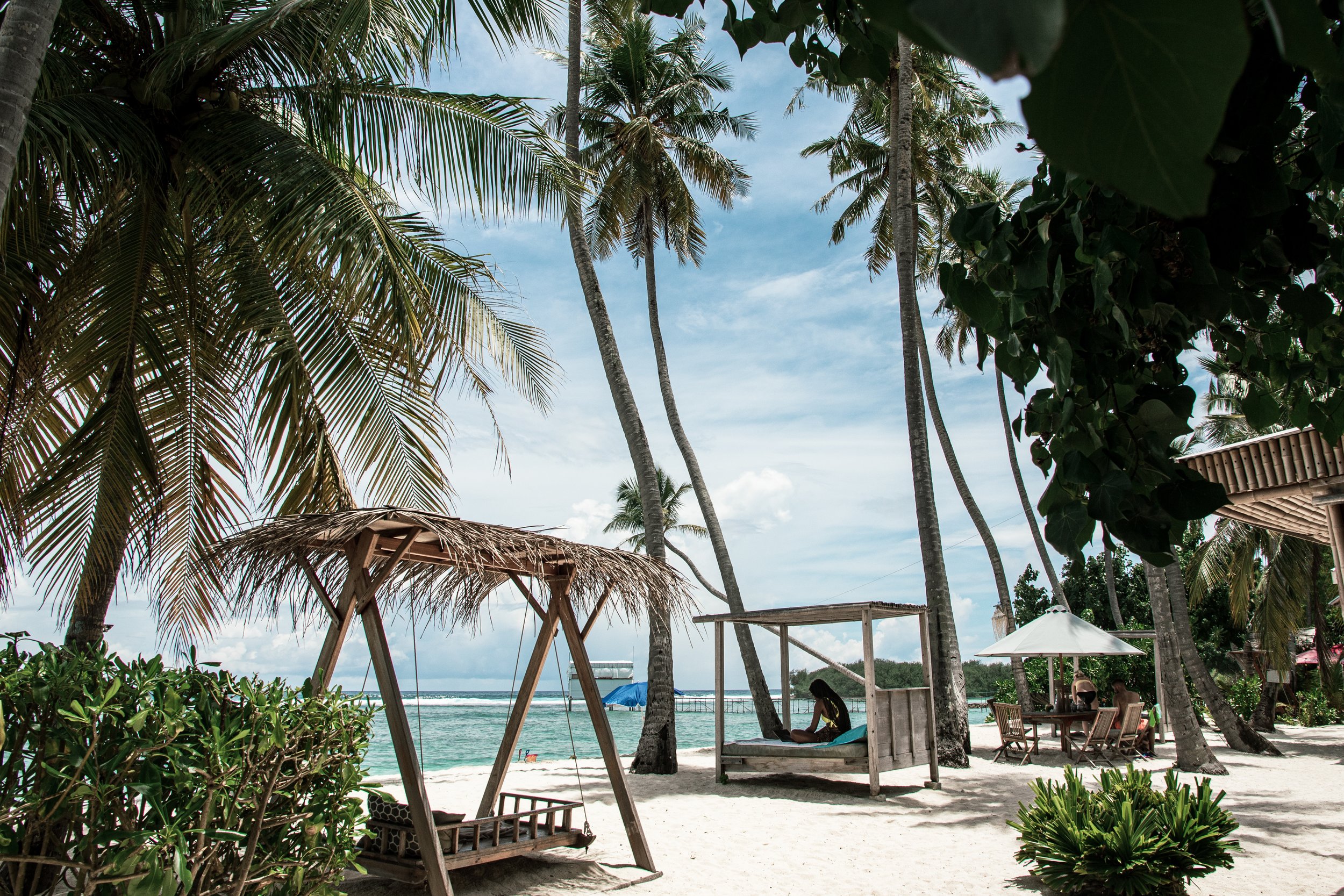 Tropical beach setting with palm trees, wooden swings, cabanas, and lounge chairs on white sand overlooking the ocean.
