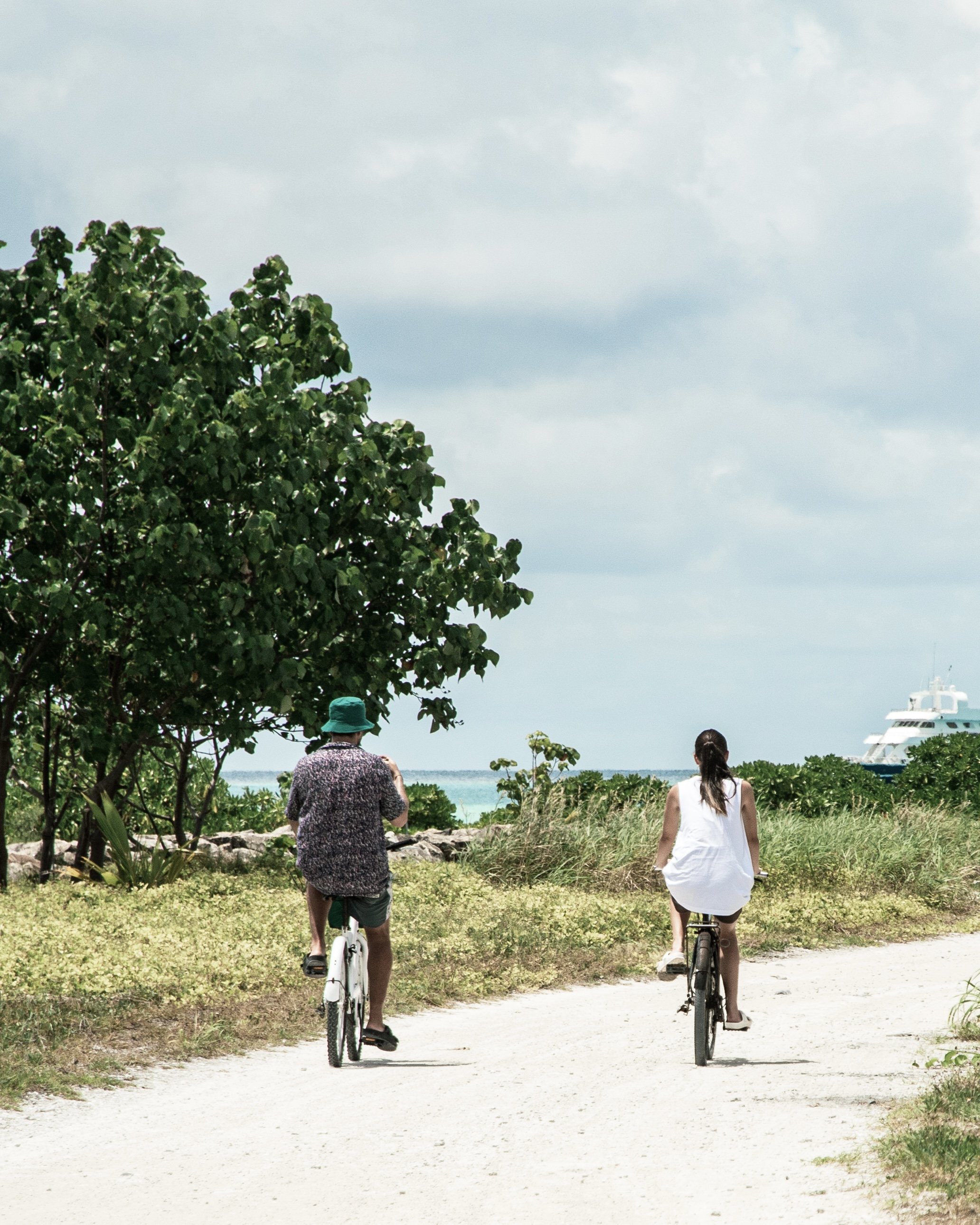 Two people cycling on a sandy path beside greenery, with a boat visible in the distance over the ocean.