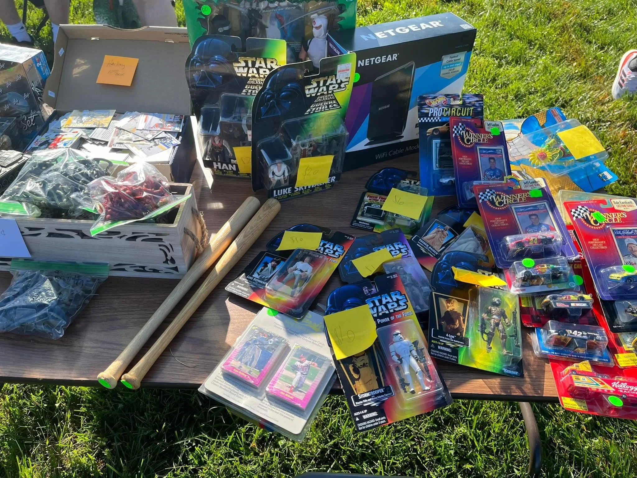 A table displaying various vintage collectibles and toys, including Star Wars action figures, die-cast cars, and packaged toy sets, with price tags and packaging. There's also a box of carded collectible figures, wooden sticks, a box of toy accessories, and some miscellaneous small items.