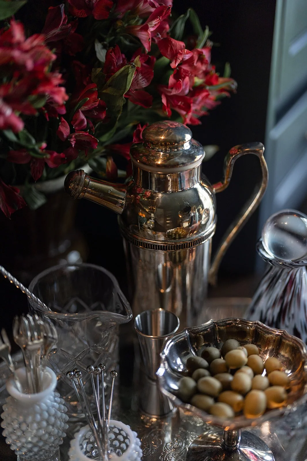A silver ice cream scoop and a silver teapot are placed on a table with a bowl of white beans and various glassware, with pink and red flowers in the background.