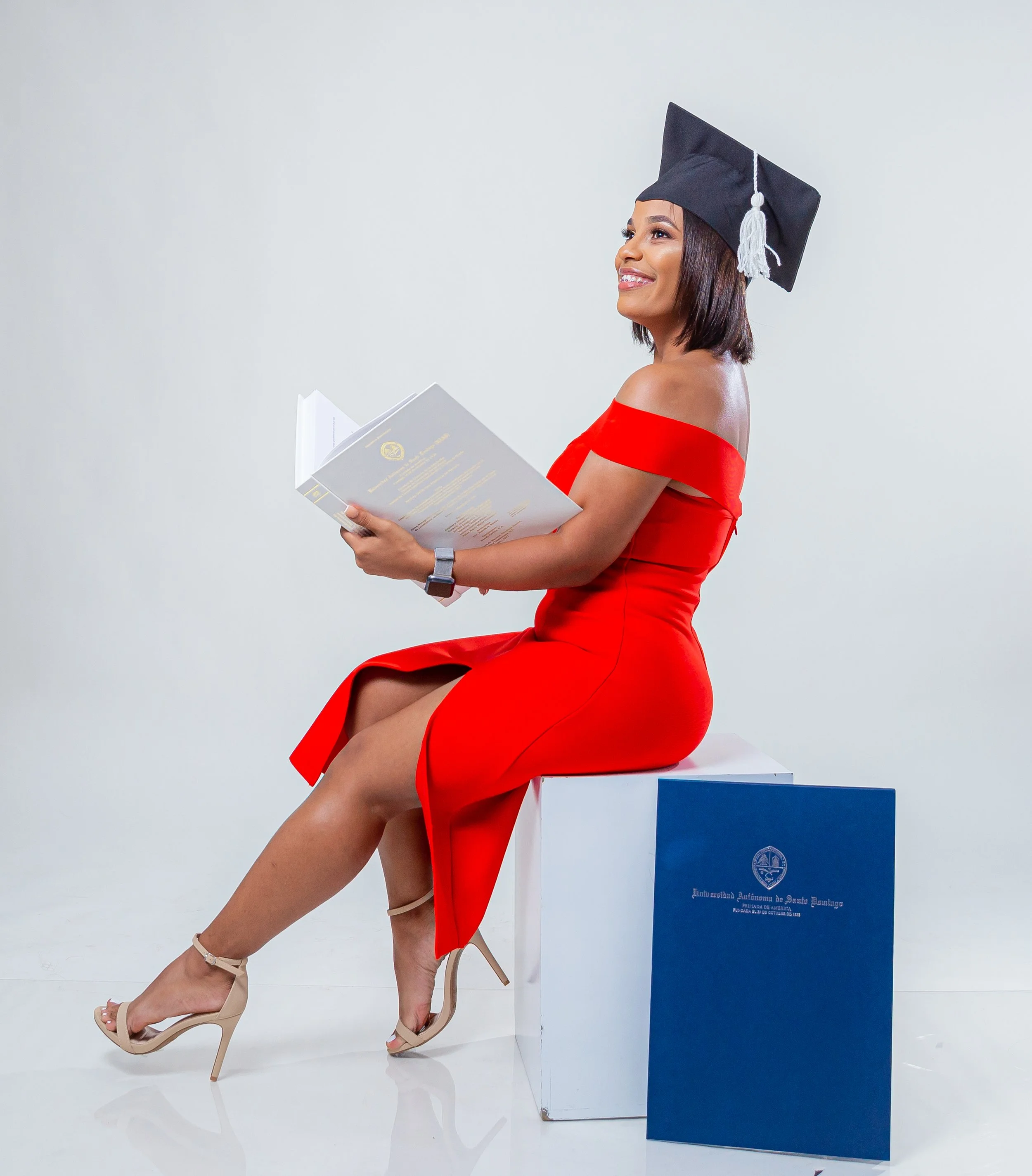 Joven mujer con toga y birrete de graduación, sentada sobre un pupitre, leyendo un documento, con un cartel en la mano, en un fondo blanco.