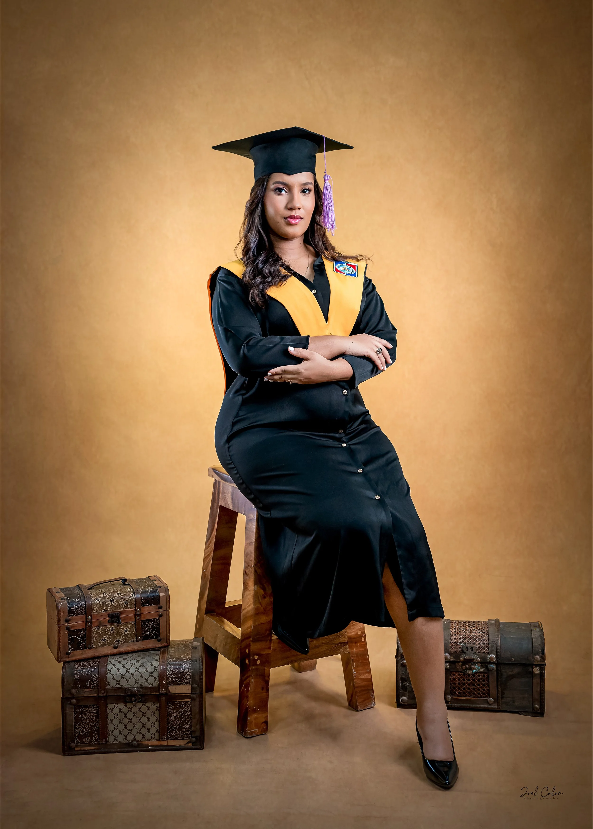Mujer vestida con toga de graduación, con birrete, sentada en una banca de madera, rodeada de dos cofres decorativos, fondo marrón
