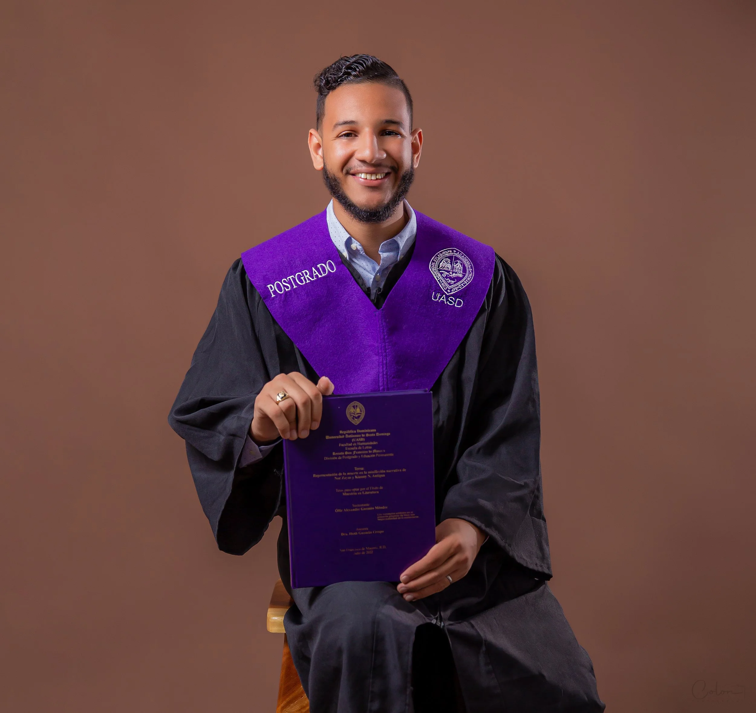 Joven graduado en toga con toga púrpura y túnica negra, sosteniendo su diploma y sonriendo en un fondo marrón.