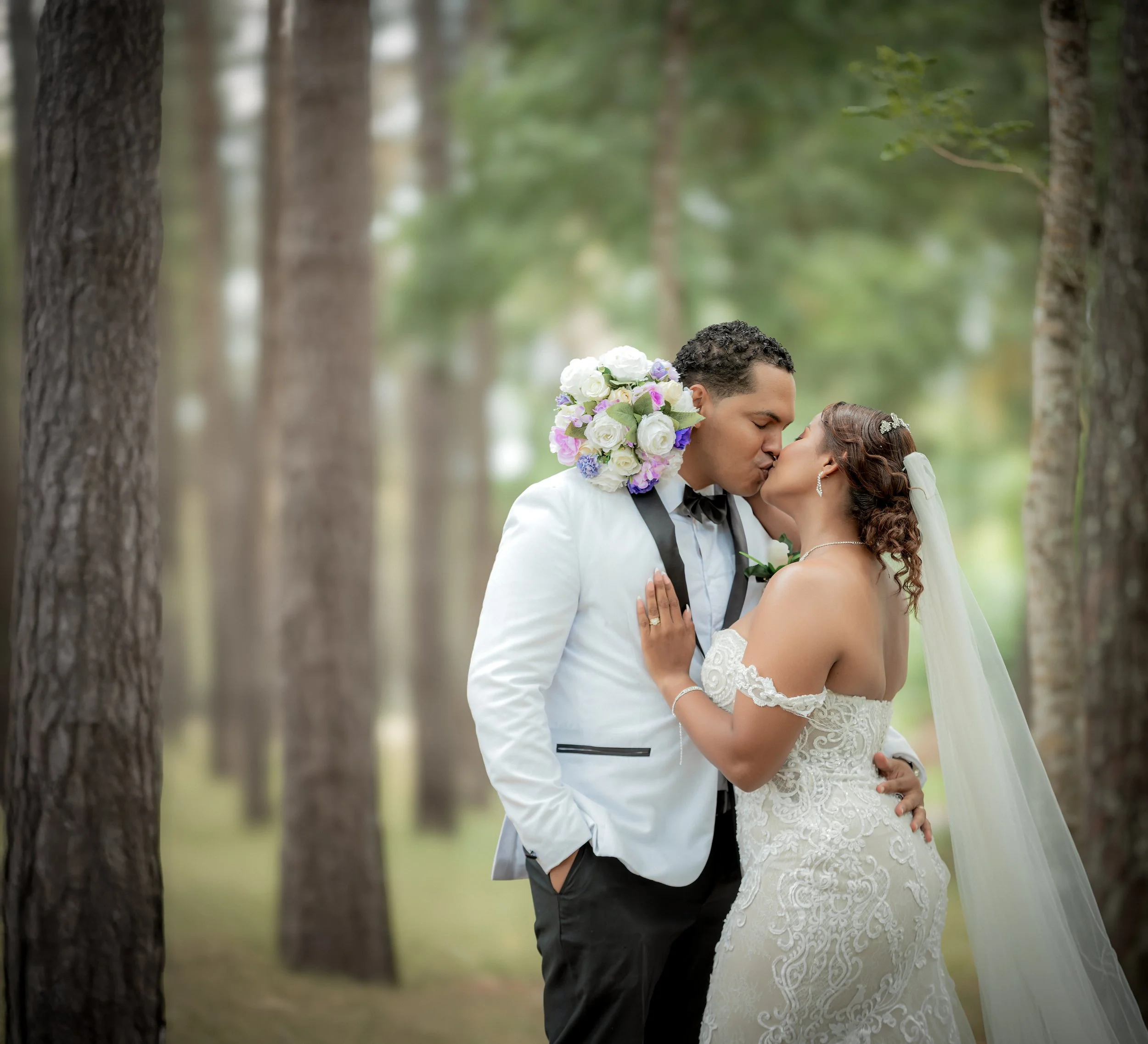 Pareja de novios besándose en un bosque, ella con vestido de novia y velo, él con traje blanco y corbata negra, rodeados de árboles