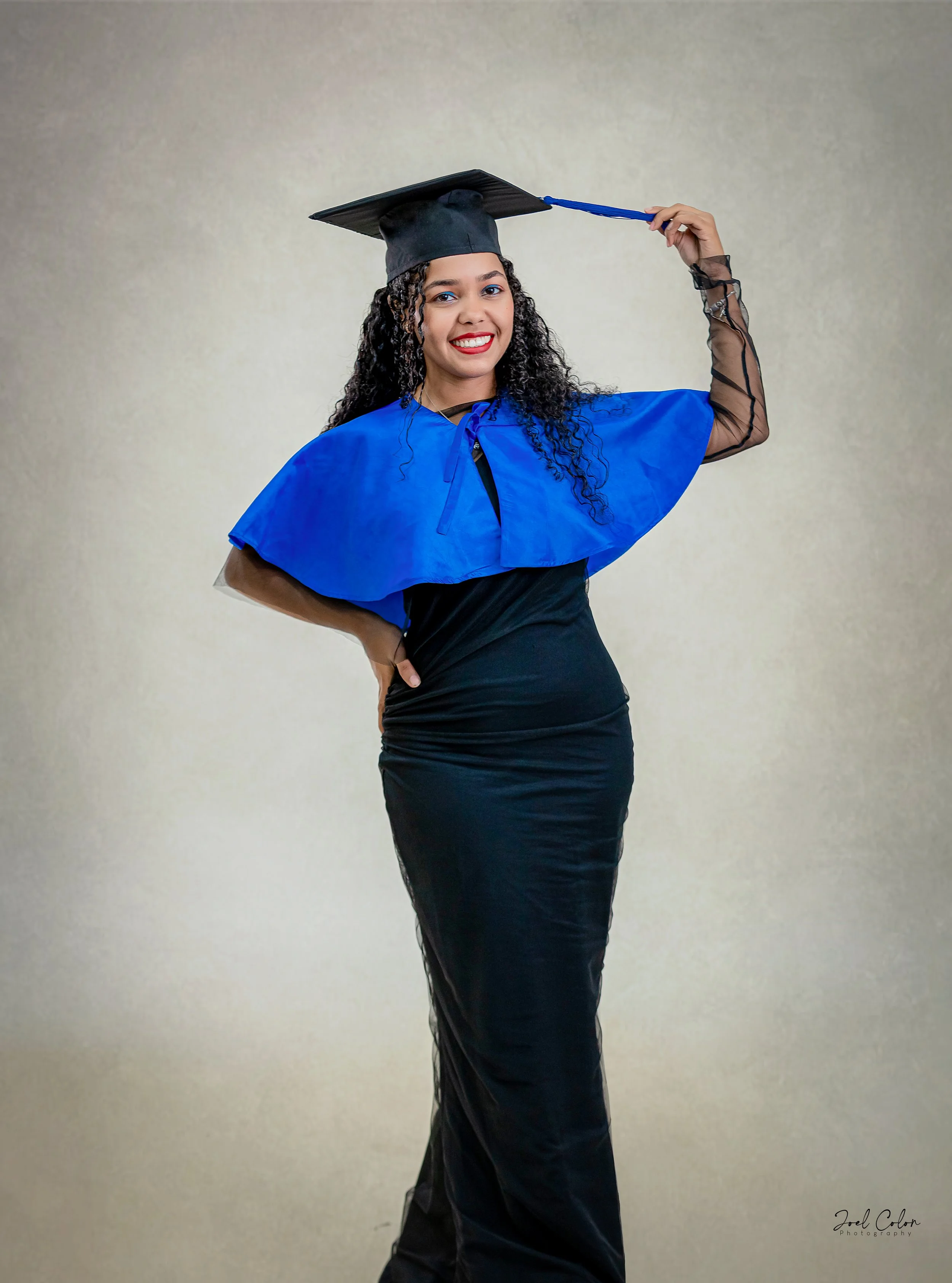 Mujer vestida con toga y birrete de graduación, sonriendo y señalando con la mano el birrete