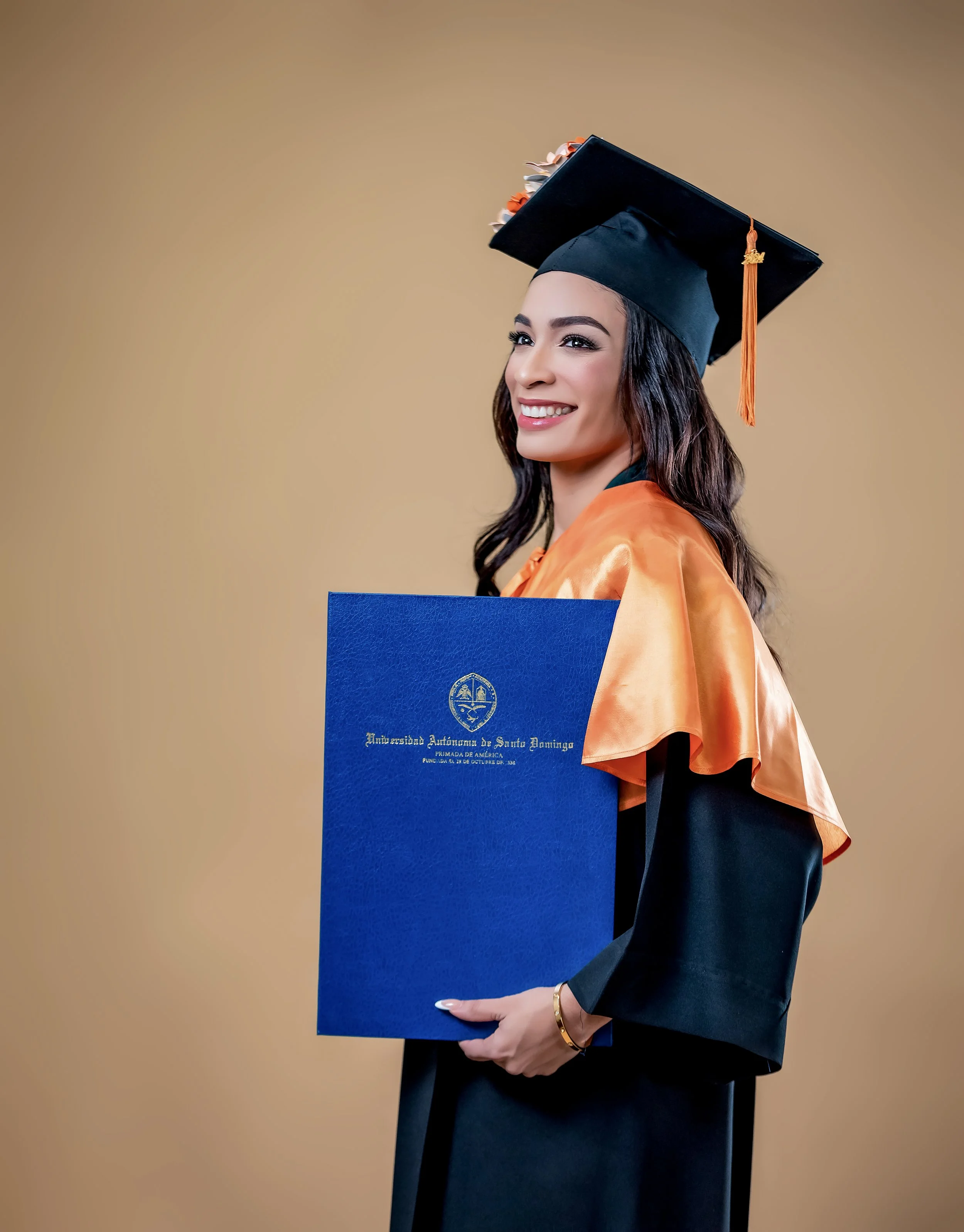 Joven graduada con vestido negro y toga, sosteniendo una diploma azul y usando un birrete negro con detalles de flores y un lazo naranja, frente a un fondo beige.