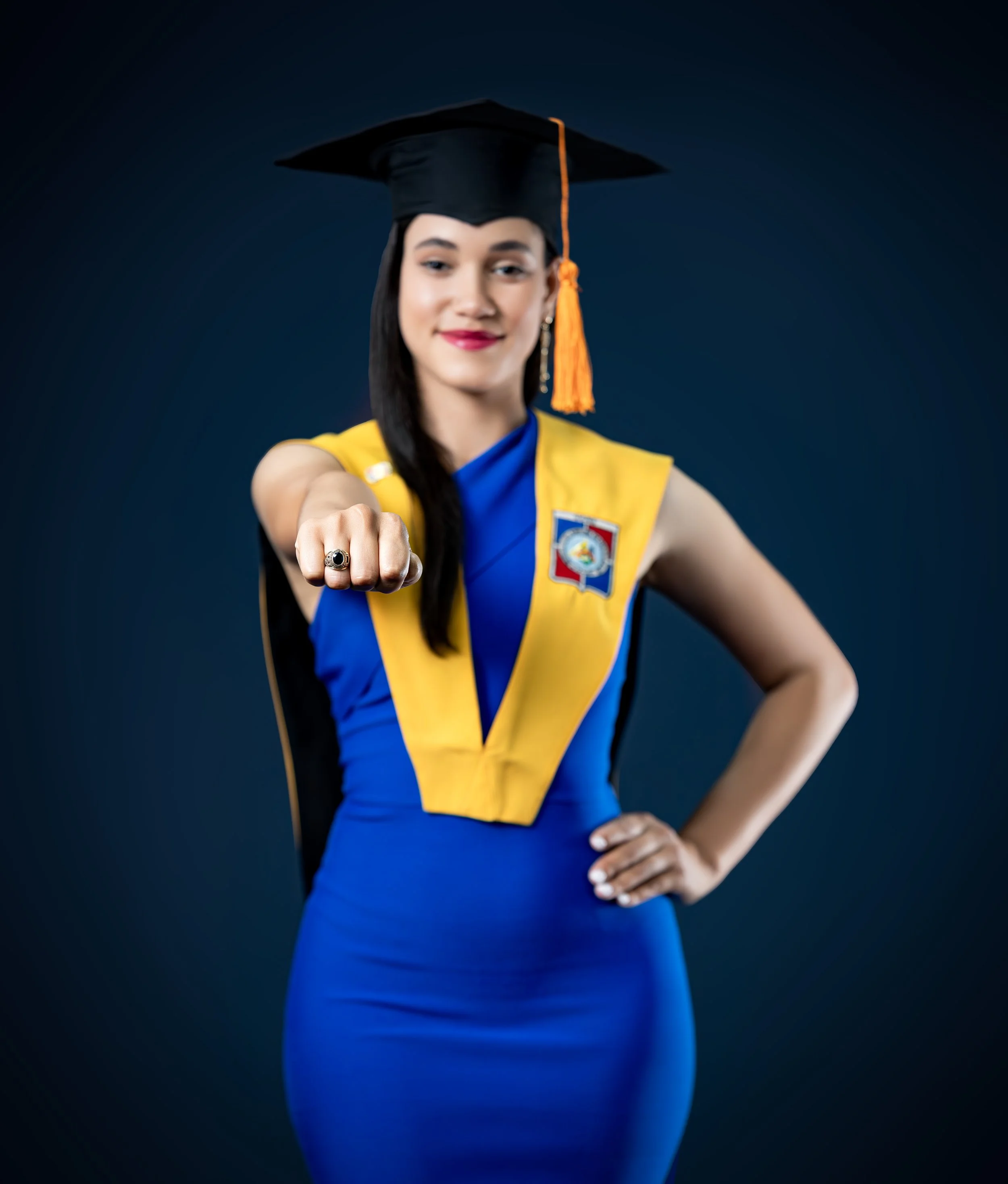 Joven mujer en toga de graduación con gorro y banda amarilla, vestido azul, posando con el puño extendido hacia adelante y una expresión confiada, con fondo oscuro.