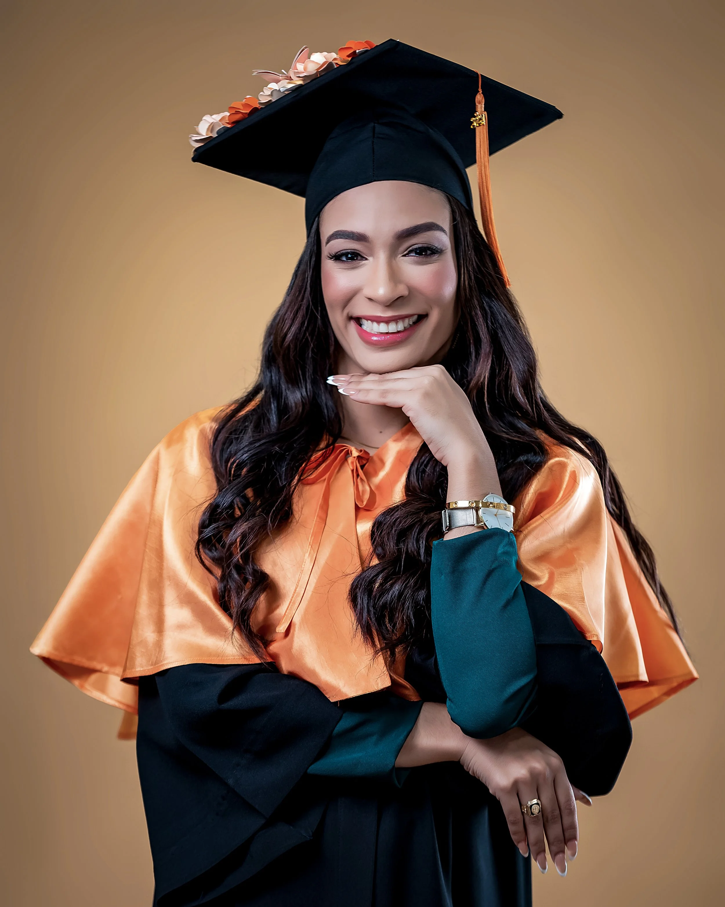 Joven mujer con graduación, vestido de graduación y birrete, sonriendo con la mano en la barbilla y fondo en color marrón.