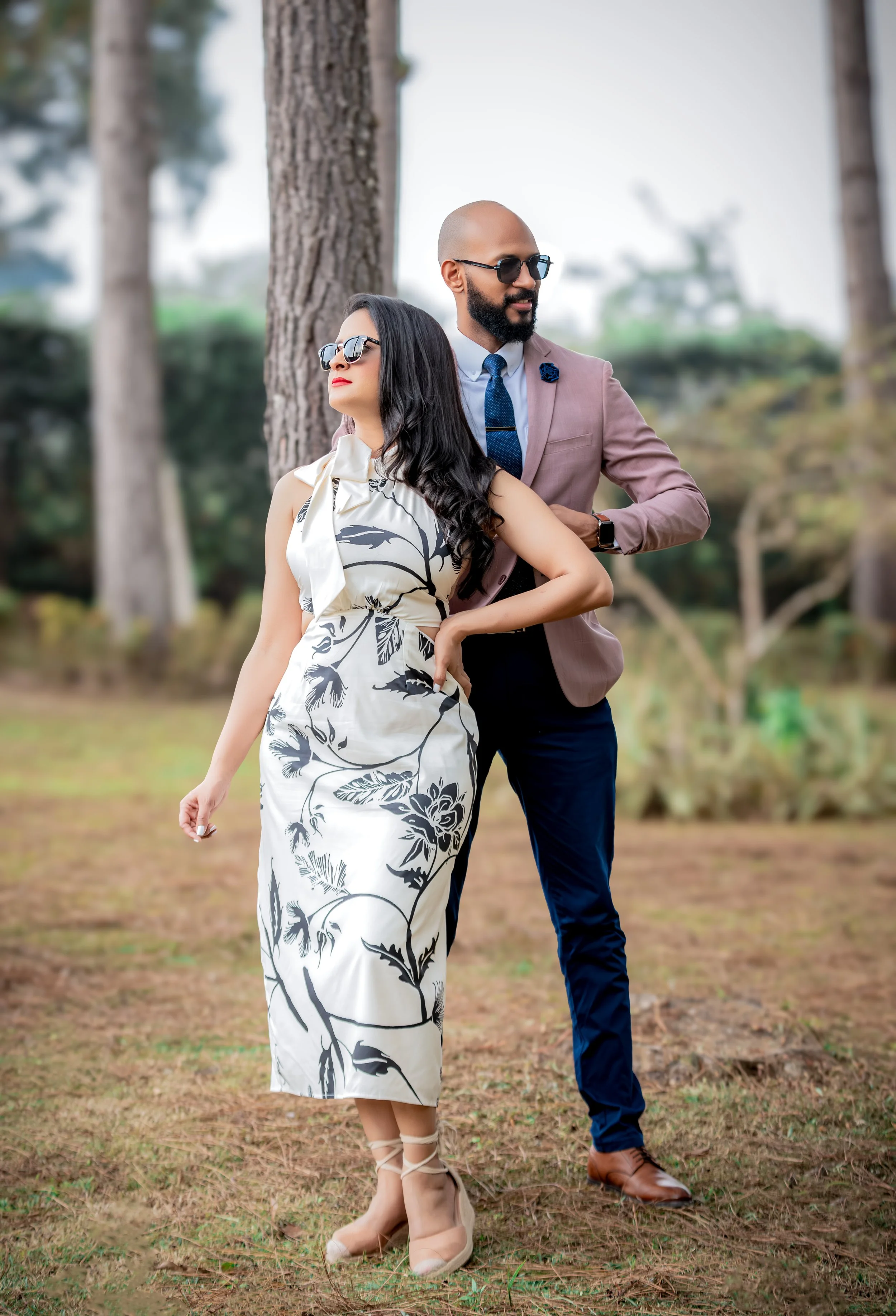 Hombre y mujer elegantemente vestida de pie en un parque con árboles, posando con actitud confiada.