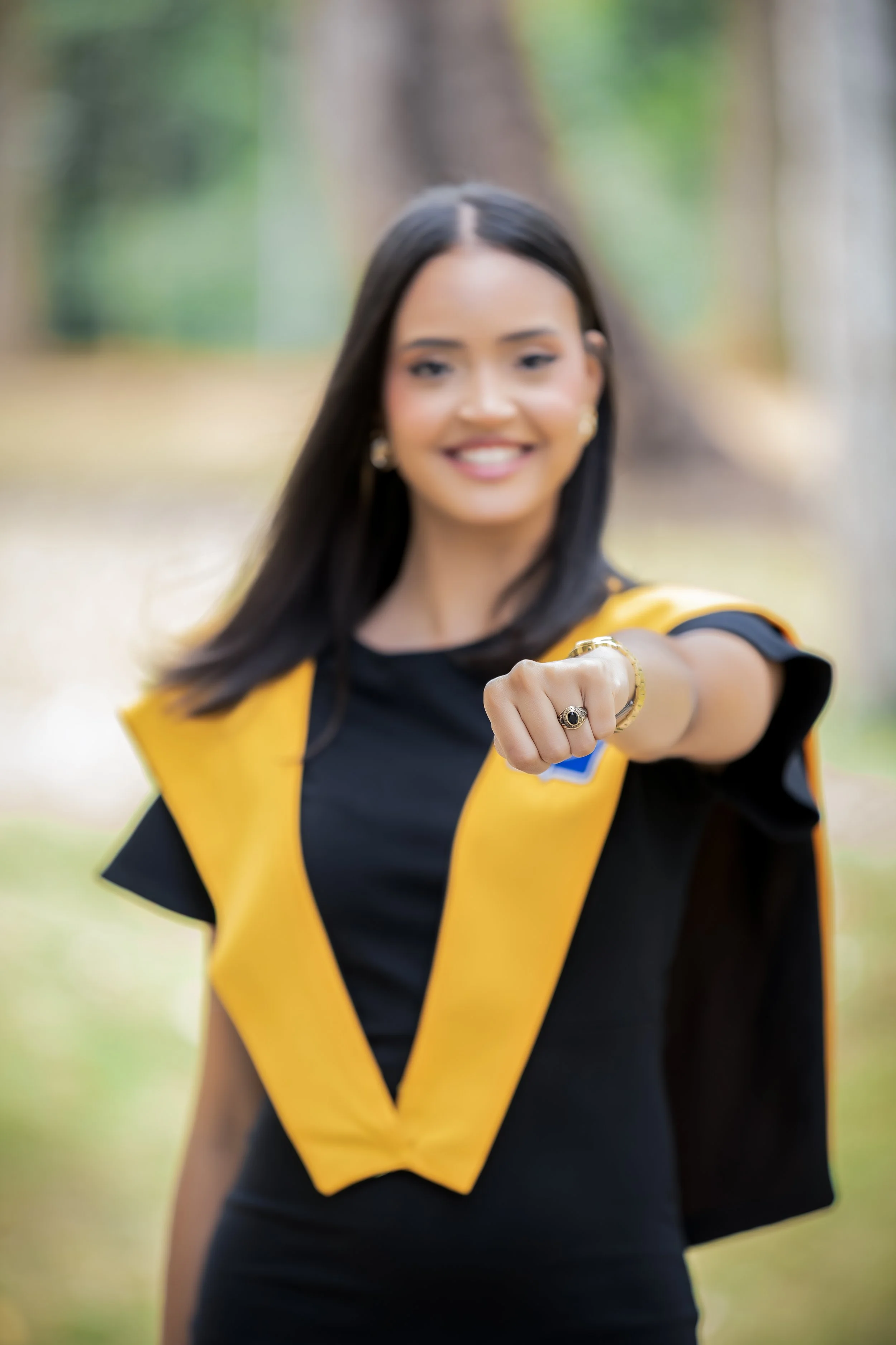 Joven mujer sonriendo, vestida con toga de graduación amarilla y negra, en un entorno al aire libre con árboles.