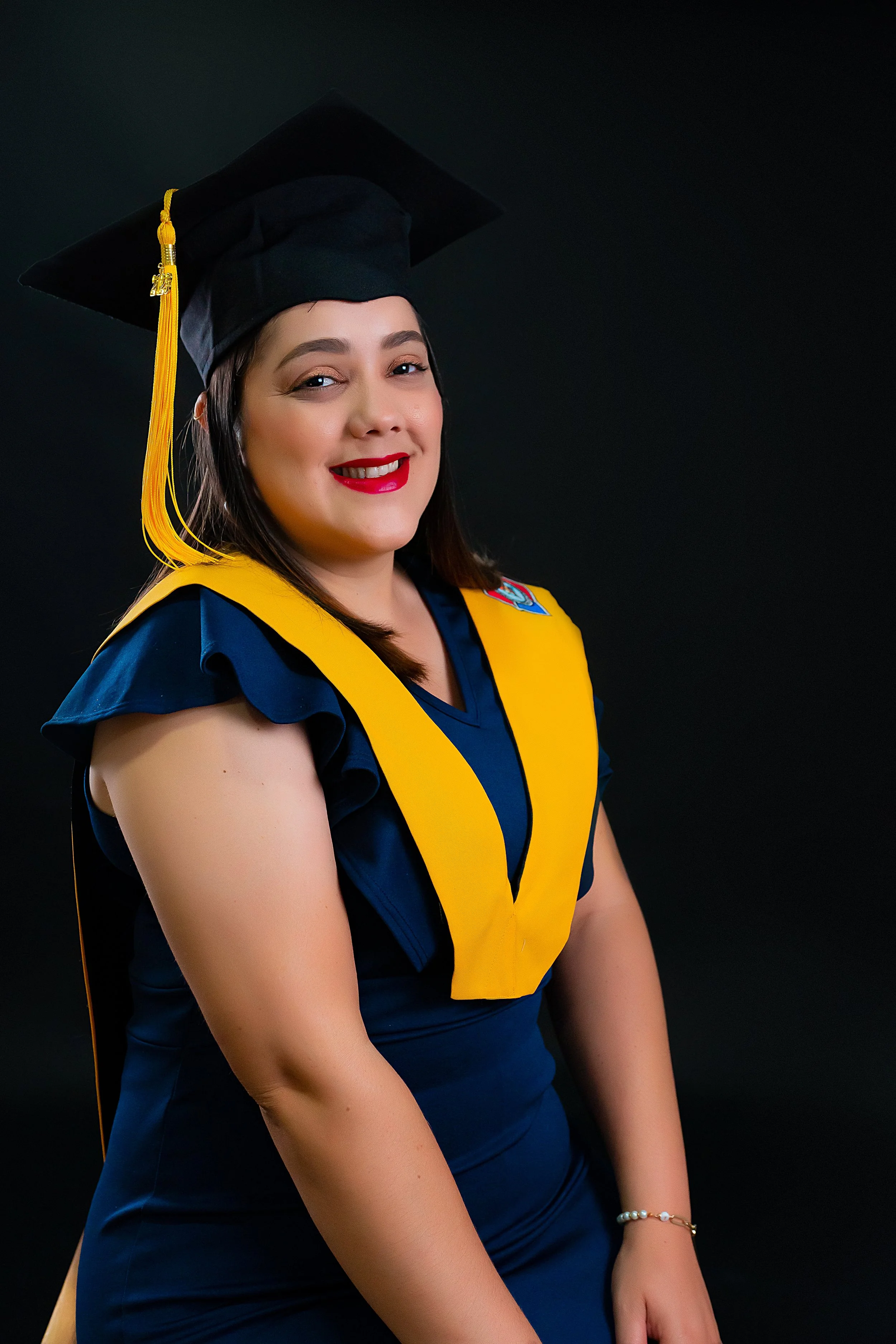 Joven mujer en toga y birrete de graduación, sonriendo, vestida con uniforme azul y con detalles en amarillo, en un fondo oscuro.