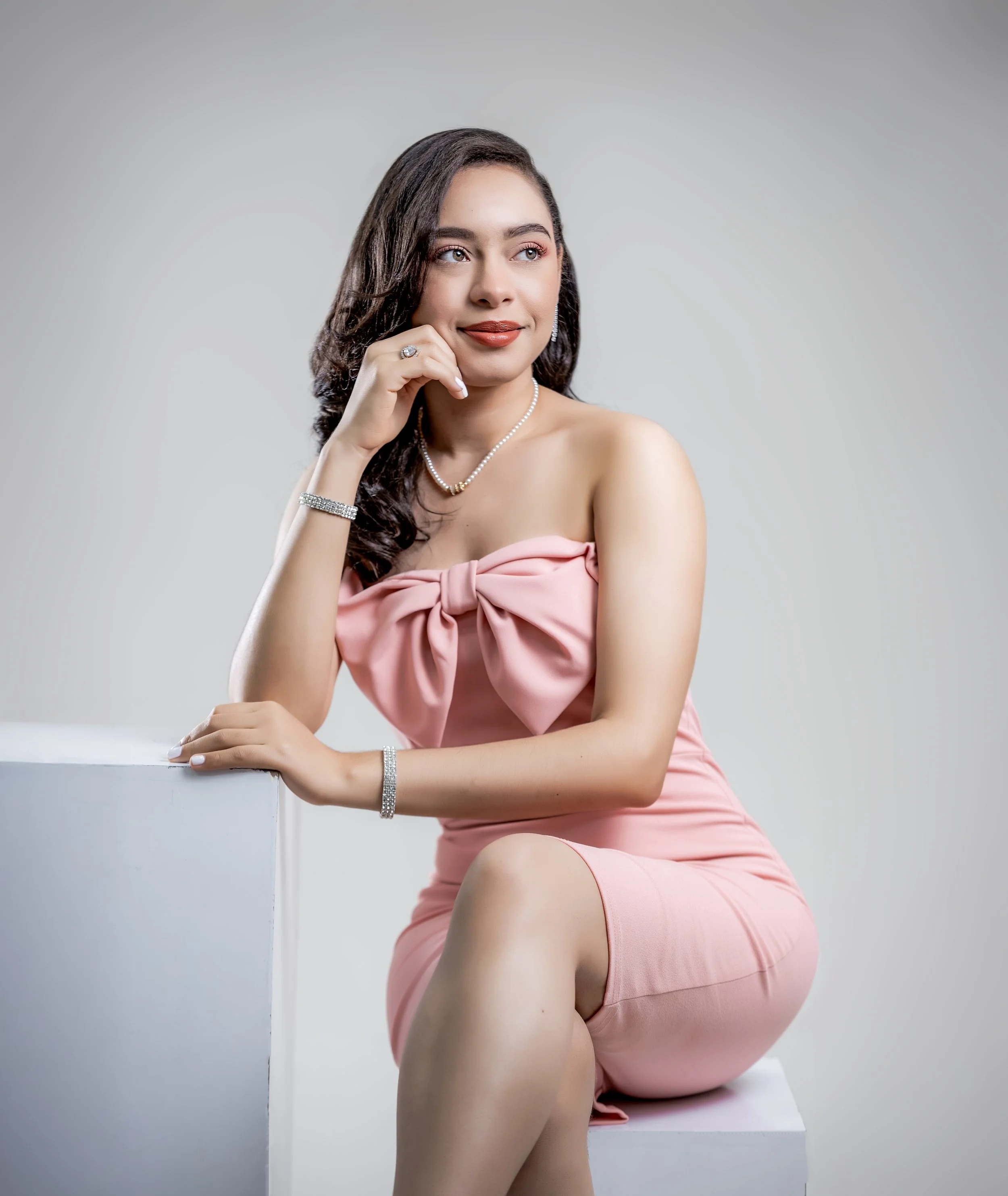 Jovencita con vestido rosa con gran lazo en el pecho, usando joyería de perlas y cristal, sentada en un banco blanco, en un fondo gris claro.