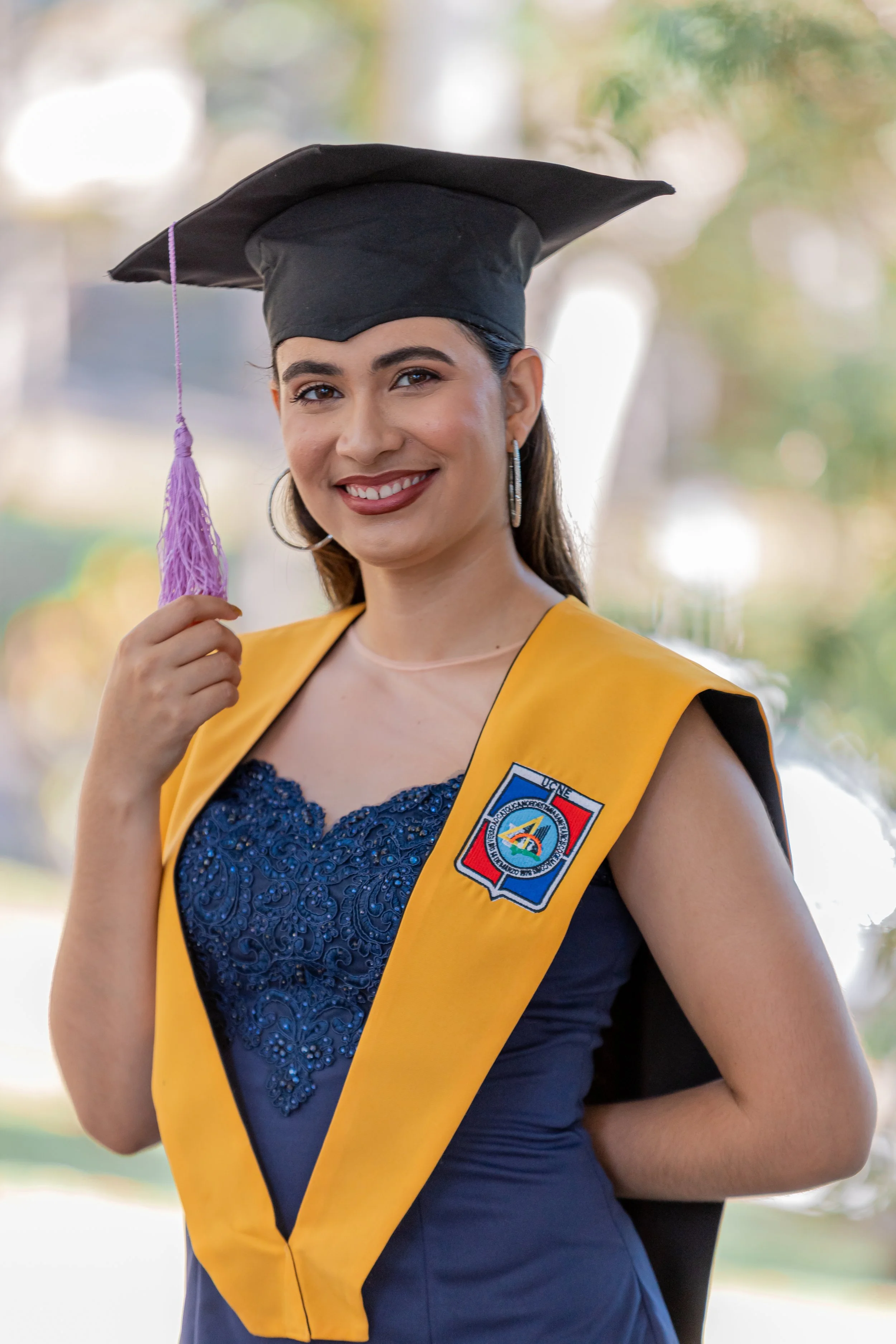 Una mujer con toga y birrete de graduación, sonriendo y mostrando la figurilla de su graduación, en un fondo borroso de árboles y cielo.
