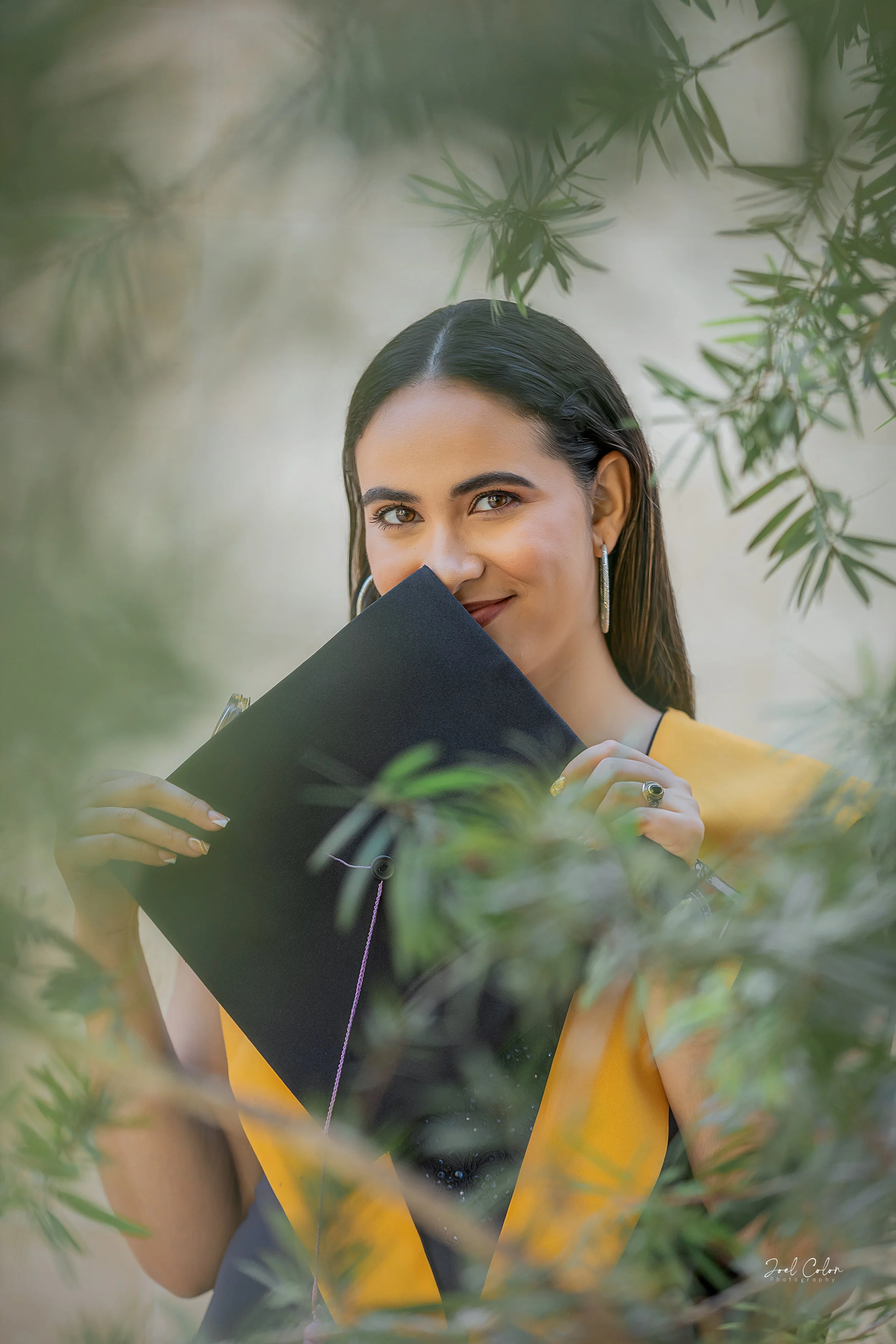 Mujer joven con vestido negro y saco amarillo, sosteniendo un portafolio negro, rodeada de vegetación, sonriendo ligeramente.