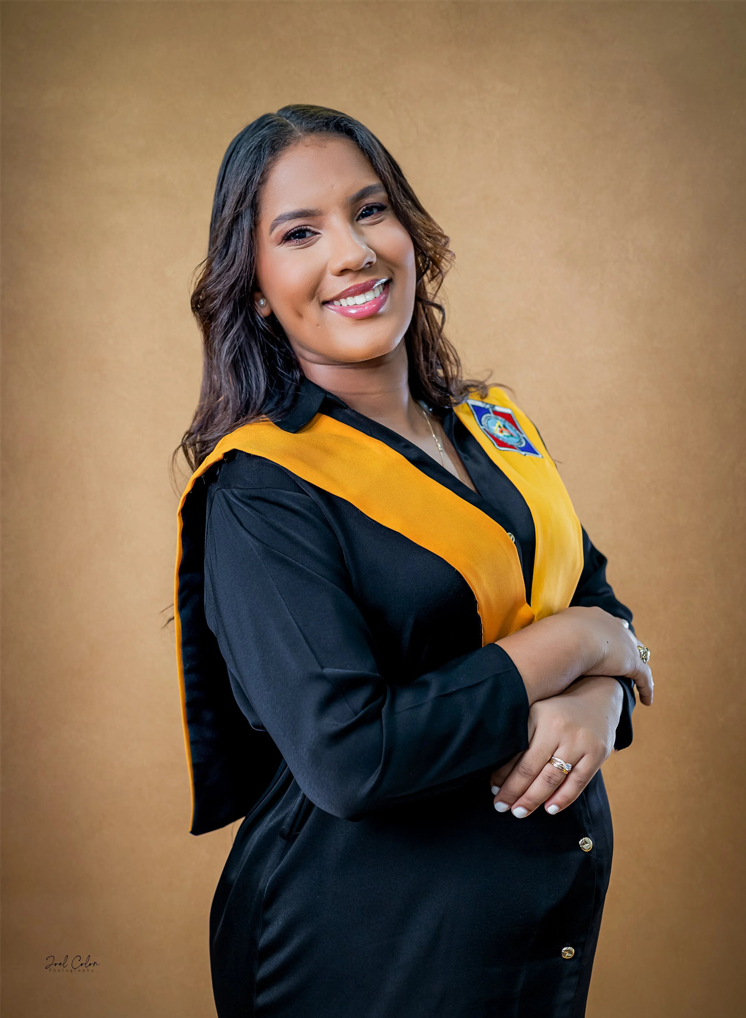 Mujer con cabello castaño oscuro y ondas, vestida con toga negra y estola amarilla, de pie con brazos cruzados, sonriendo en un fondo dorado.