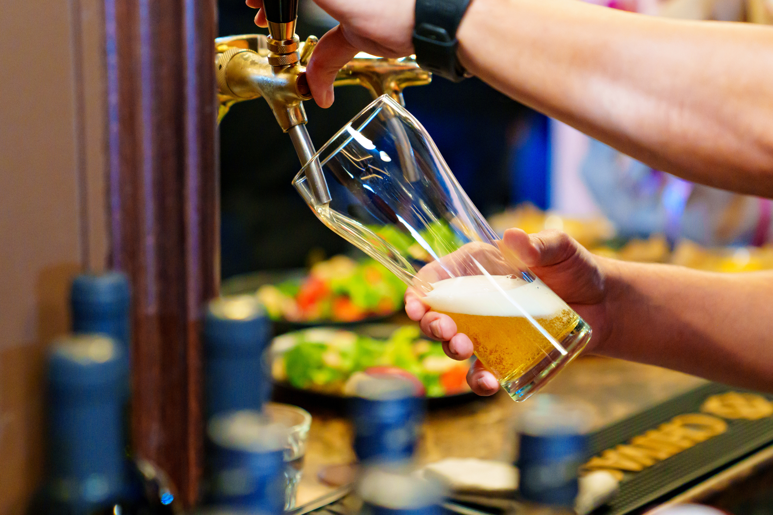 Person pouring draft beer into a glass from a tap, with bottles and a tray of food in the background.