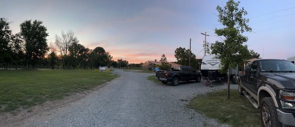 Gravel road with parked trucks and a trailer at sunset, surrounded by trees and grass.