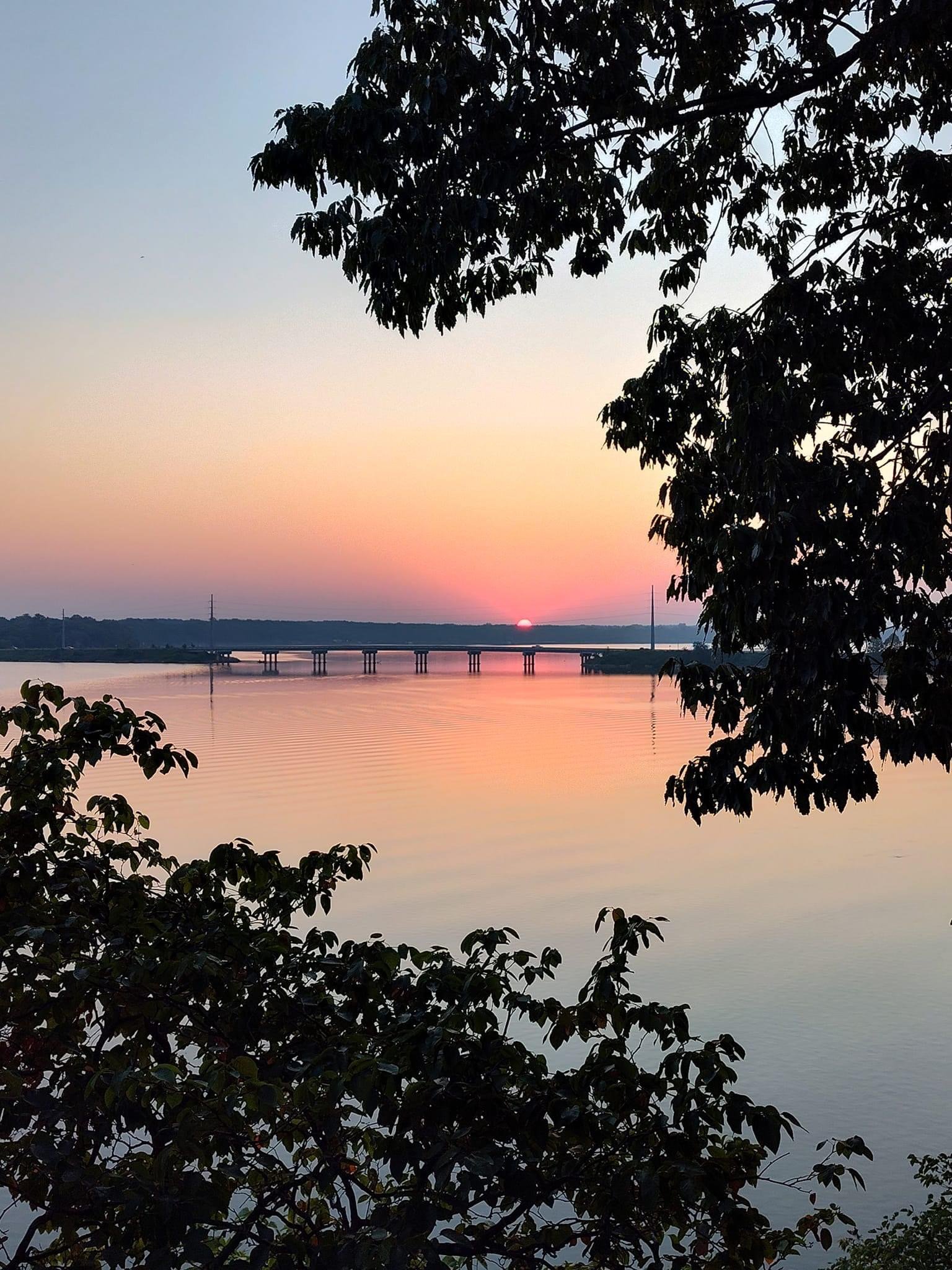 Scenic view of a sunset over a calm lake with a bridge in the distance, framed by silhouetted tree branches.