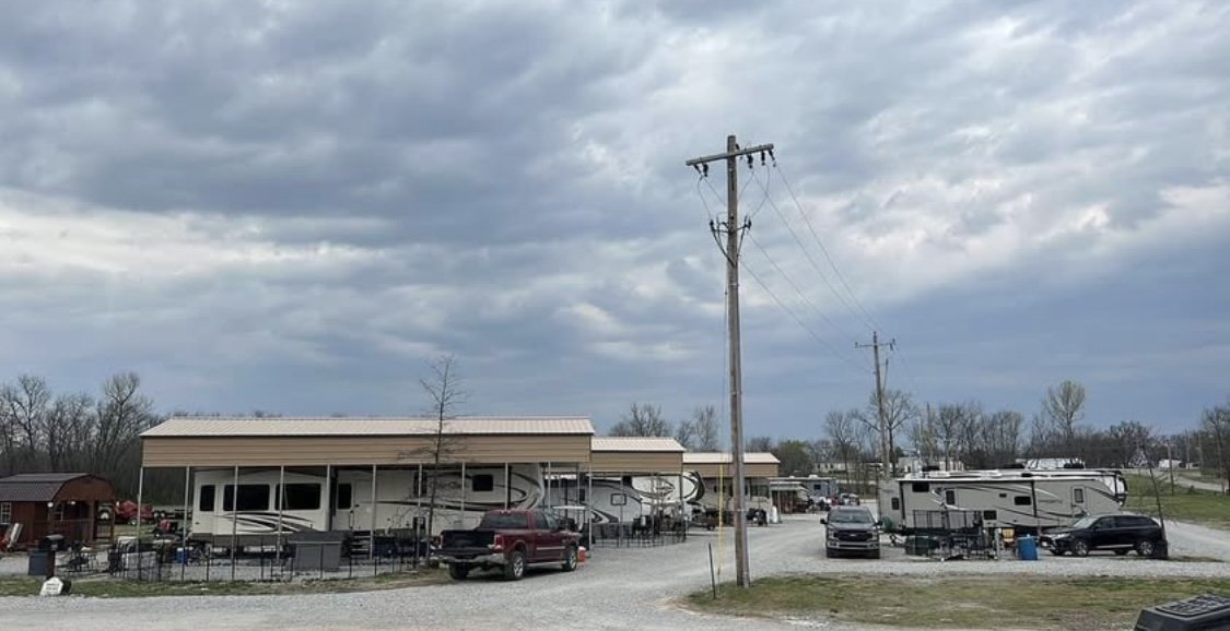 A rural RV park with multiple RVs parked under shelters, a gravel road, parked trucks, and cloudy sky above.