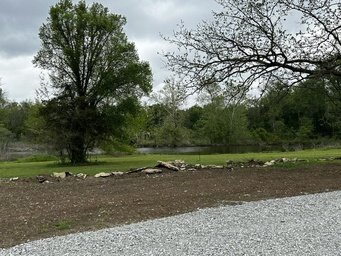Gravel road and grassy field with trees and a river in the background under a cloudy sky.
